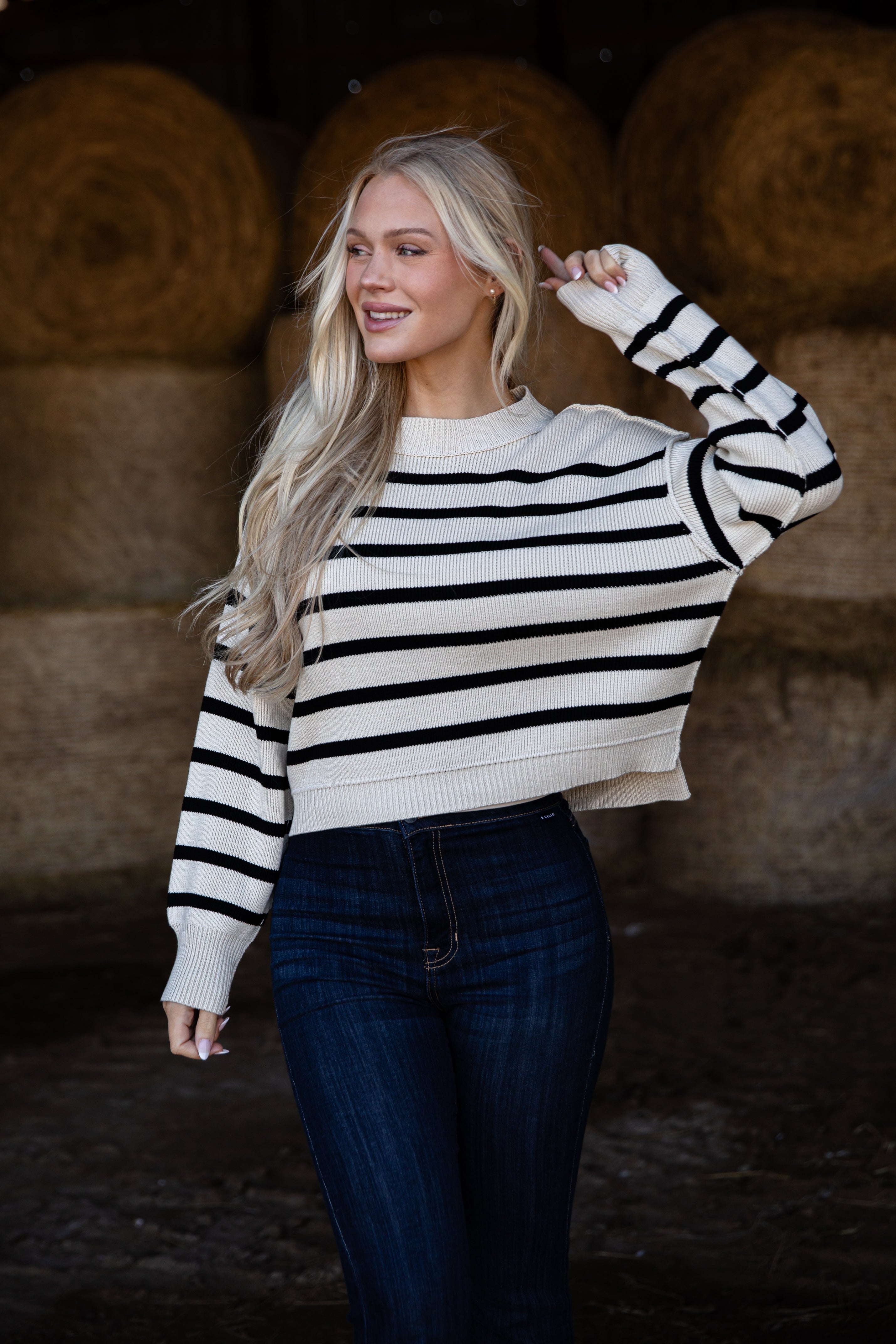 Woman wearing a black and white striped sweater and blue jeans in front of hay bales.