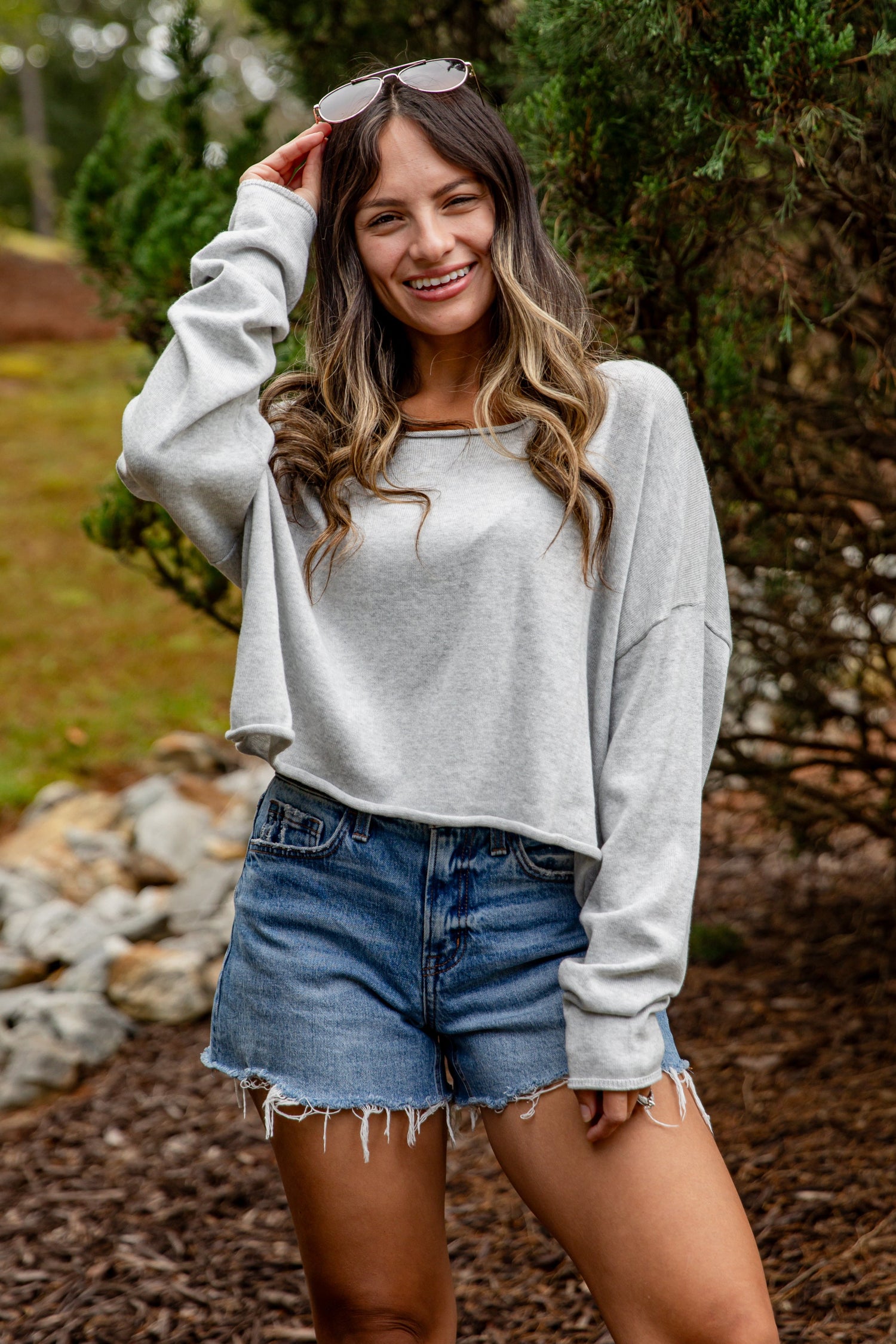 Woman wearing a gray sweatshirt and denim shorts standing outdoors with greenery in the background