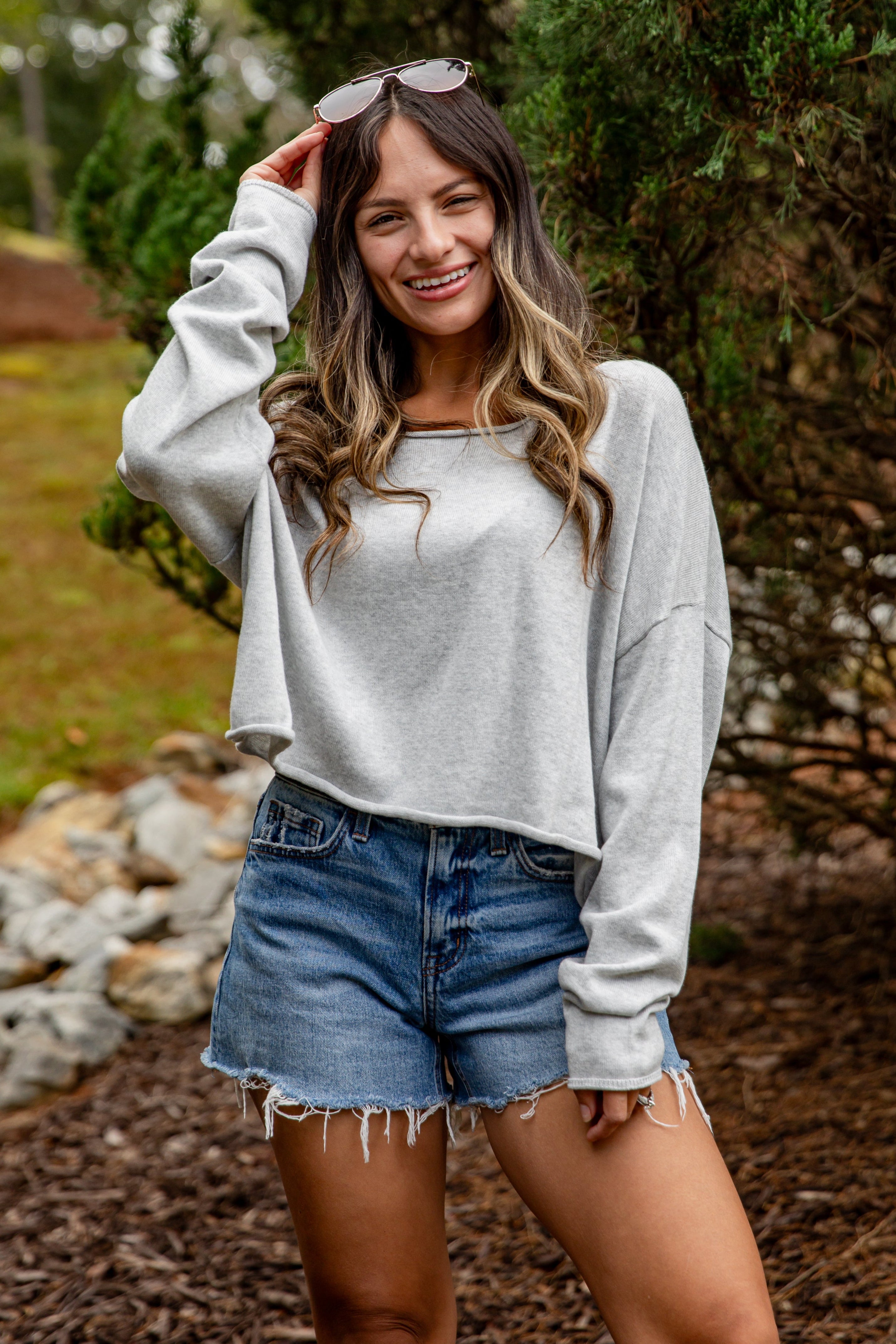 Woman wearing a gray sweatshirt and denim shorts standing outdoors with greenery in the background