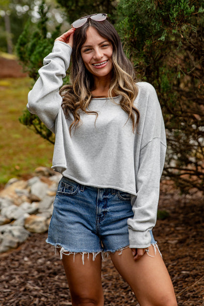 Woman wearing a gray sweatshirt and denim shorts standing outdoors with greenery in the background