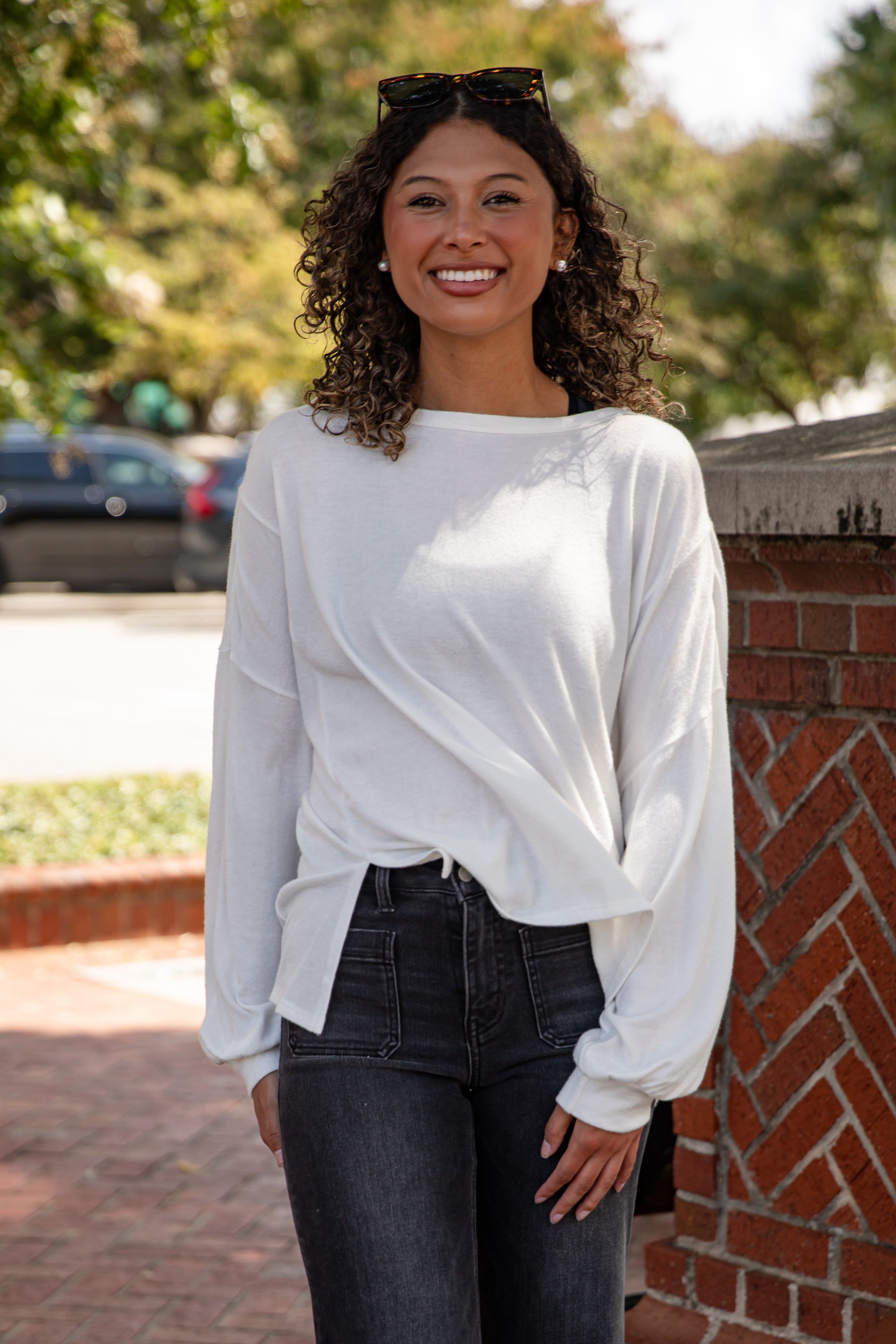 Woman wearing a white blouse and dark jeans standing outdoors with trees and a brick wall in the background.