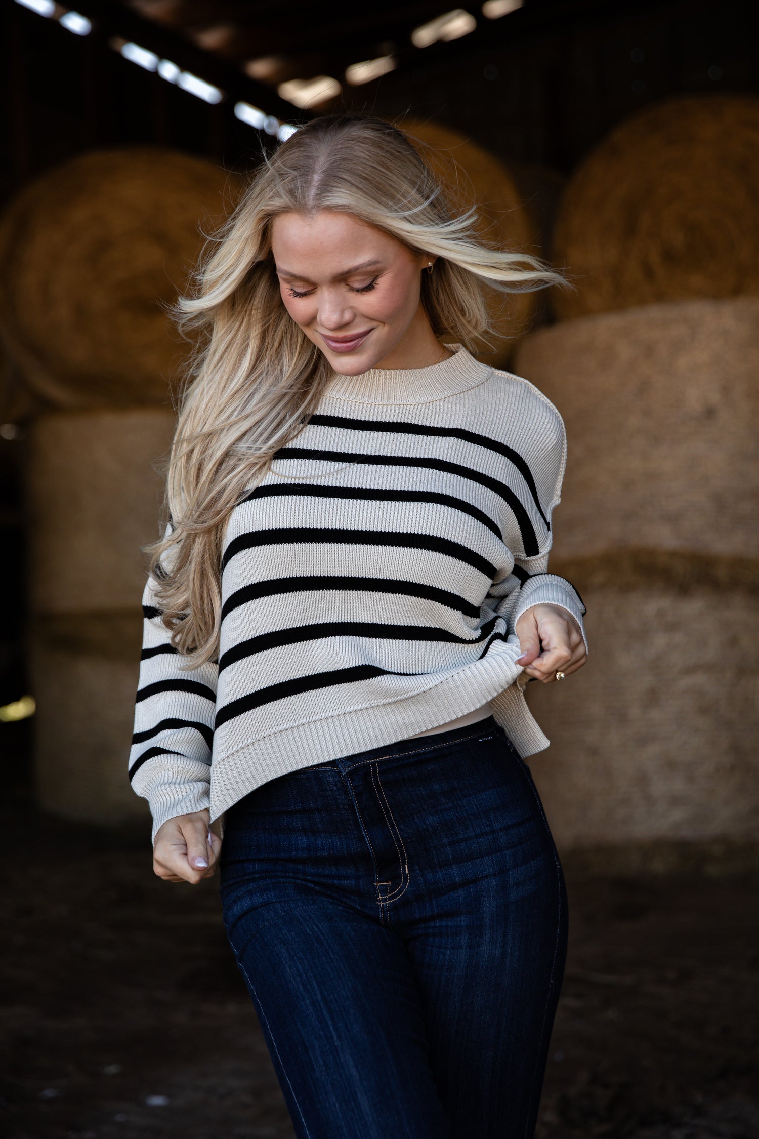 Woman wearing a black and white striped sweater and dark jeans in a barn setting with hay bales.