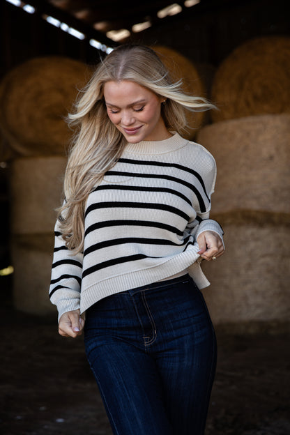 Woman wearing a black and white striped sweater and dark jeans in a barn setting with hay bales.