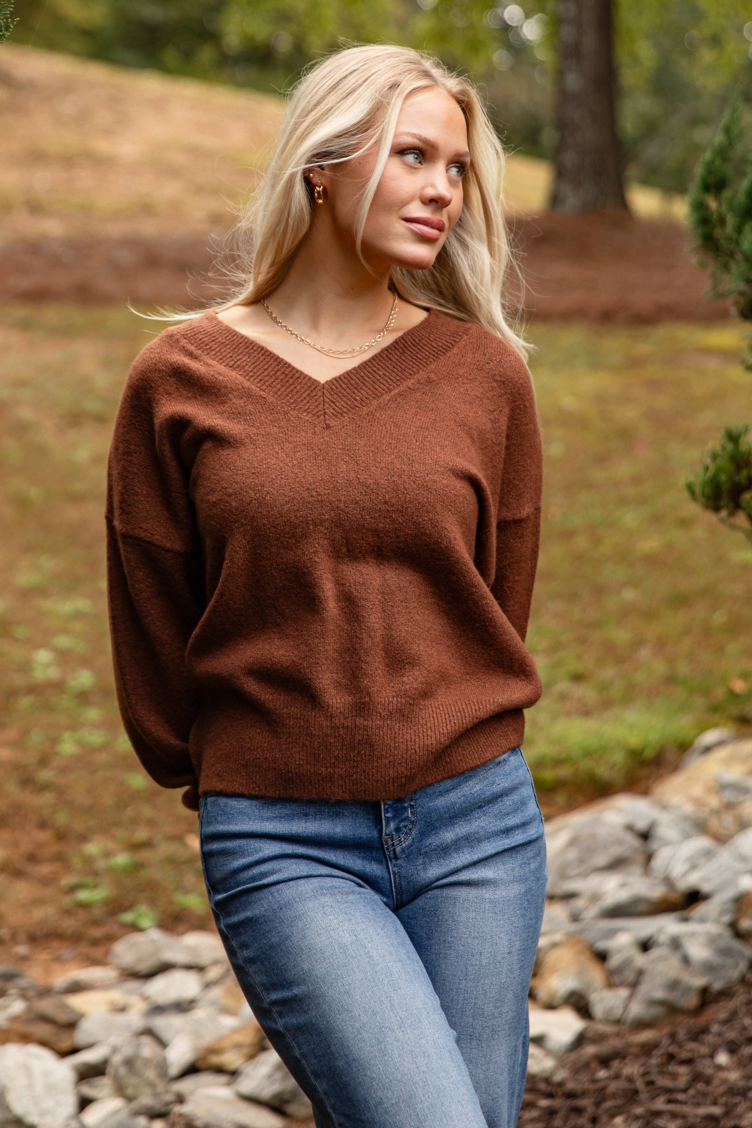 Woman wearing a brown sweater and blue jeans standing outdoors with trees and rocks in the background