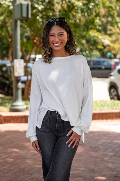 Woman wearing a white sweater and dark jeans standing on a sidewalk with trees and cars in the background.