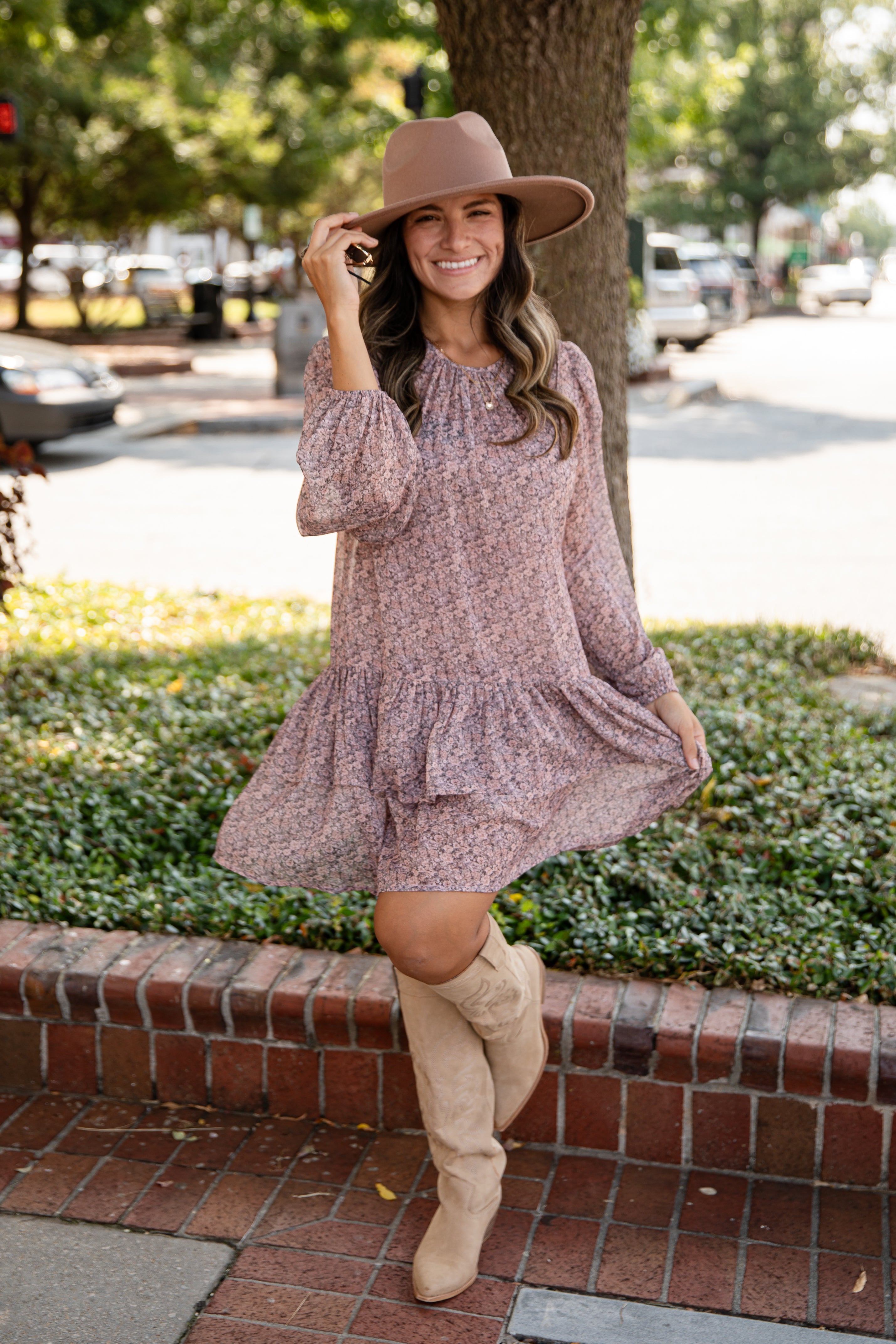 Woman in a floral dress and beige boots standing on a sidewalk with trees and cars in the background.