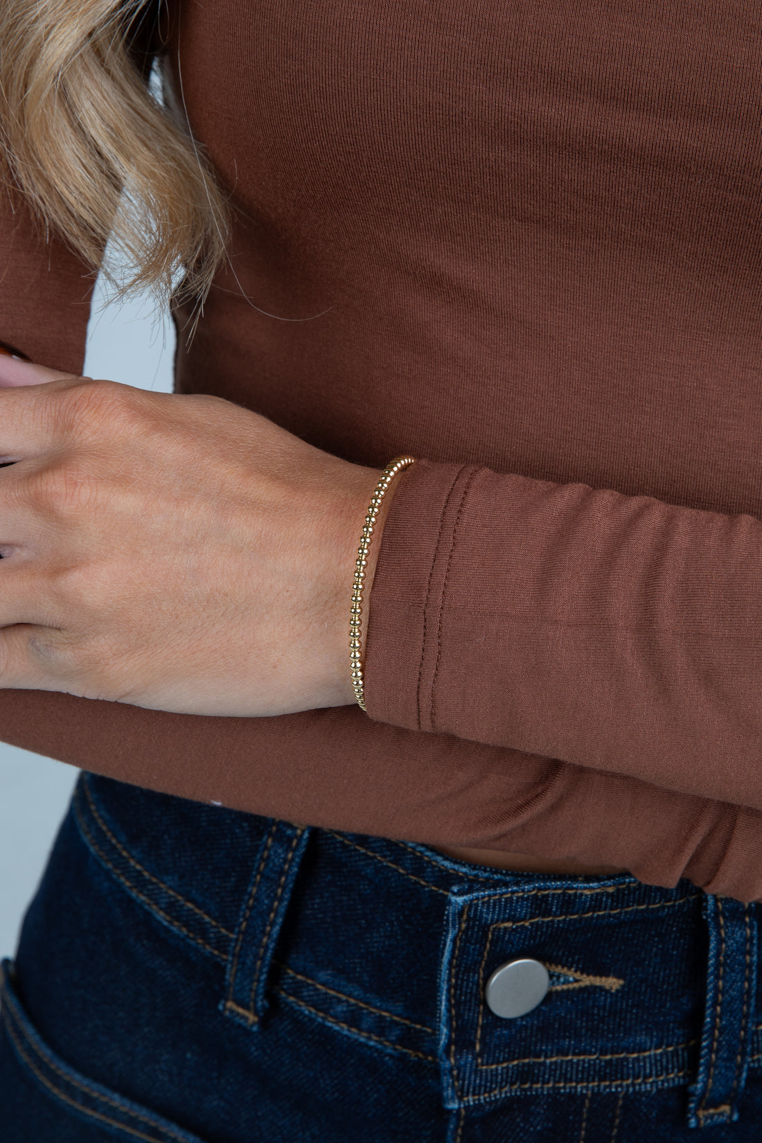 Close-up of a person wearing a brown long-sleeve shirt and blue jeans with a gold bracelet.