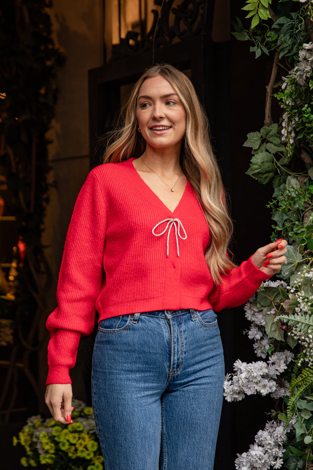 Woman wearing a red sweater with a white bow design and blue jeans, standing outdoors with plants in the background.