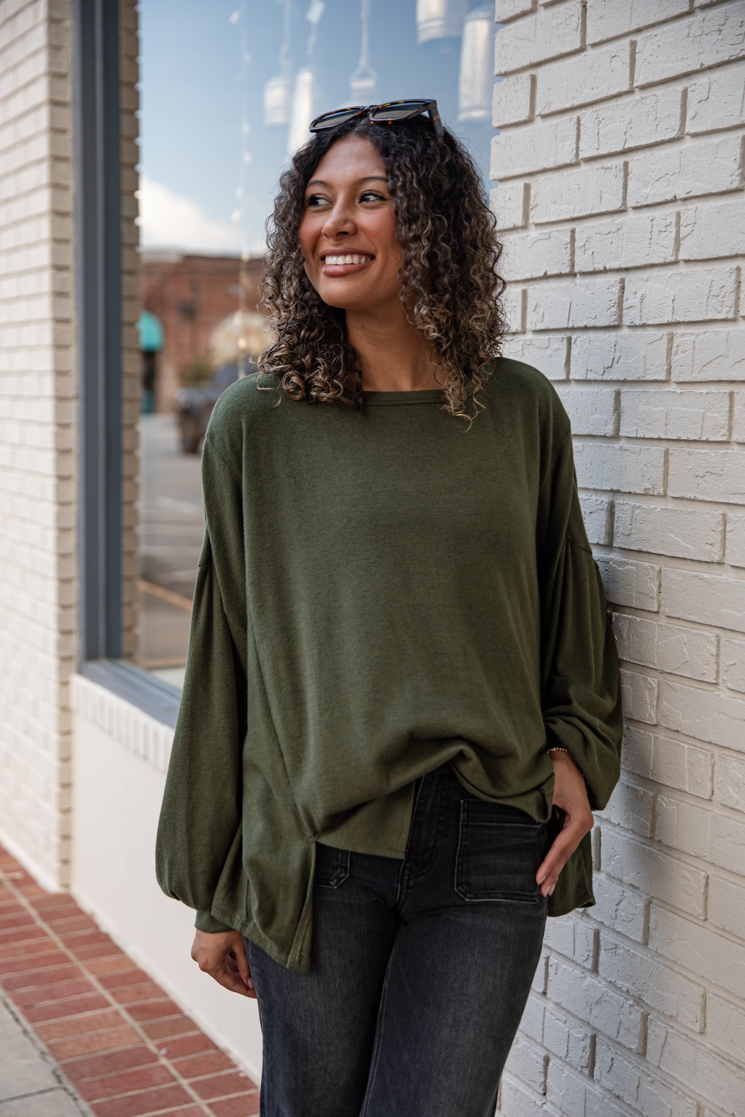 Woman wearing an olive green long-sleeve top and dark jeans standing against a brick wall.