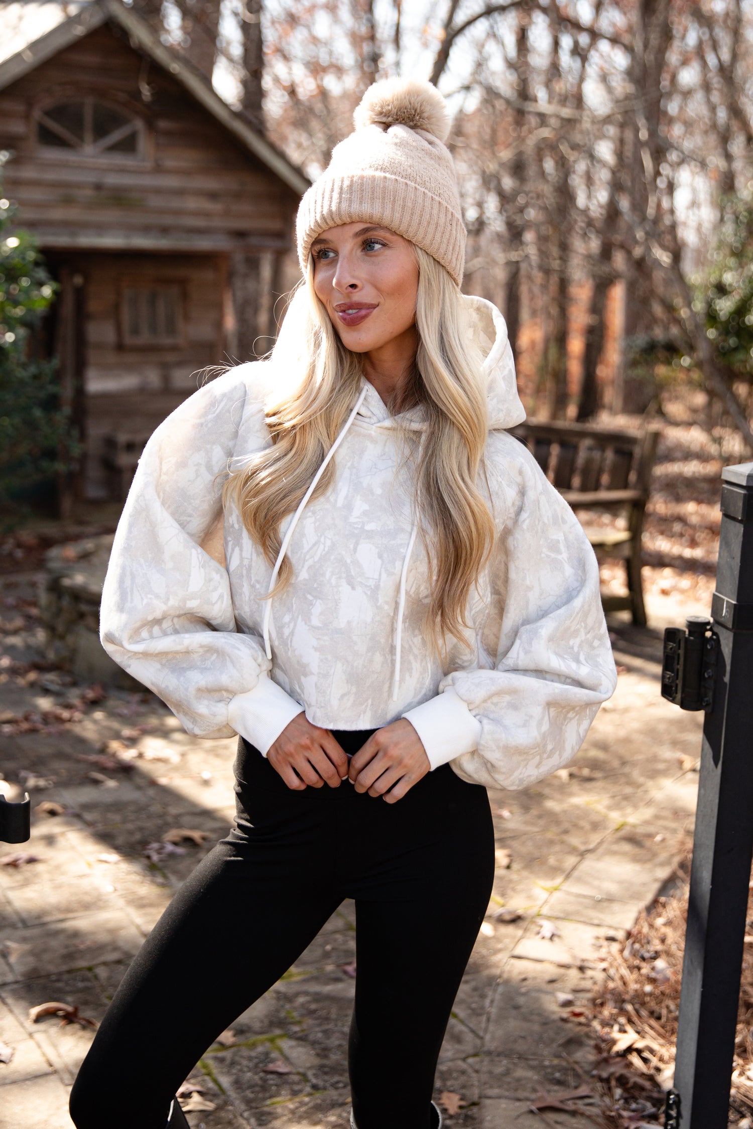 Woman wearing a white hoodie and black pants outdoors with a wooden cabin in the background