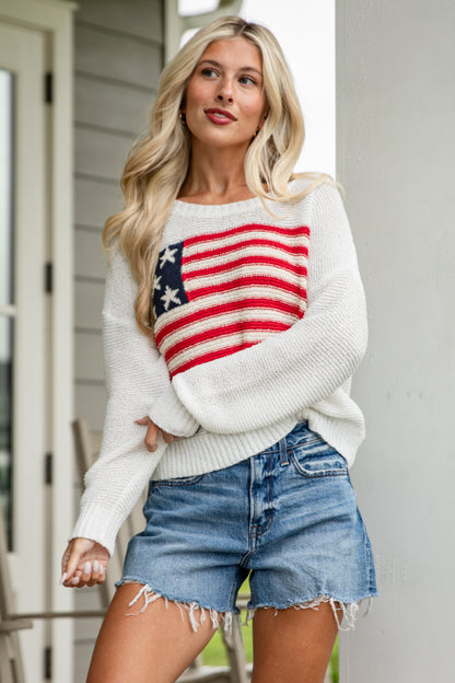 Woman wearing a white sweater with an American flag design on a porch.