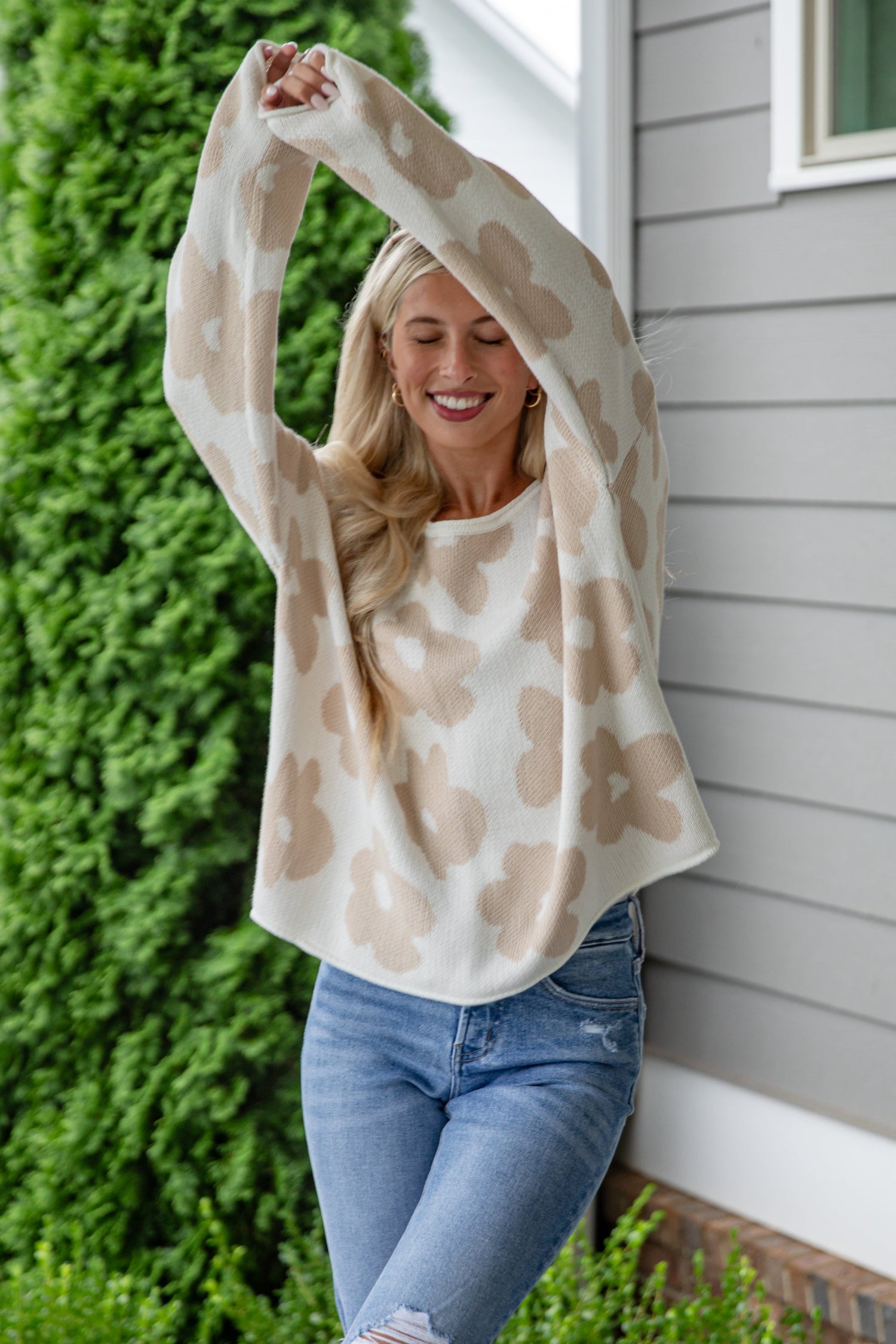 Woman in a patterned top and jeans standing on a wooden deck with greenery and a house in the background.