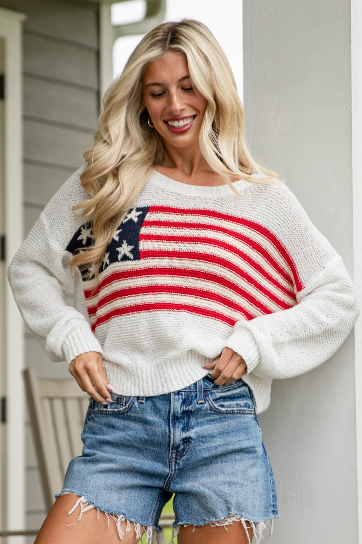 Woman wearing a white sweater with an American flag design and denim shorts on a porch.