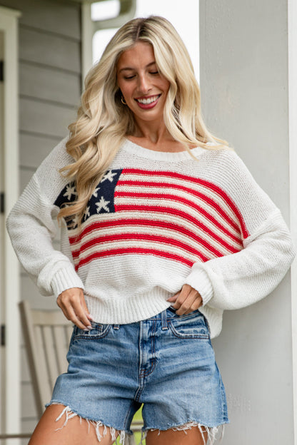 Woman wearing a white sweater with an American flag design and denim shorts on a porch.