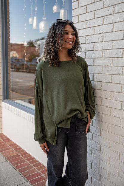 Woman wearing an olive green sweater and dark jeans leaning against a brick wall.