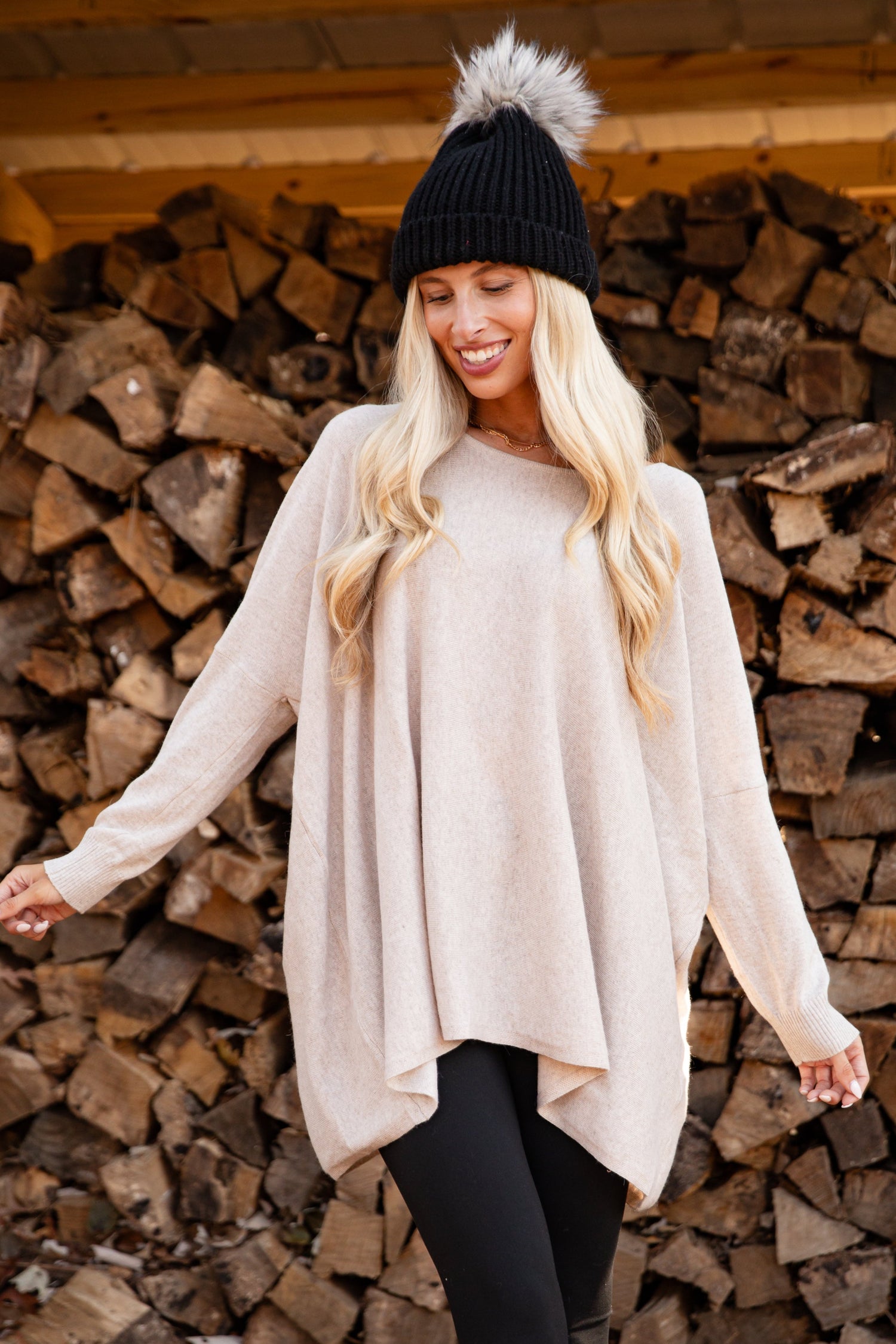 Woman in a beige sweater and black leggings standing in front of stacked wood.