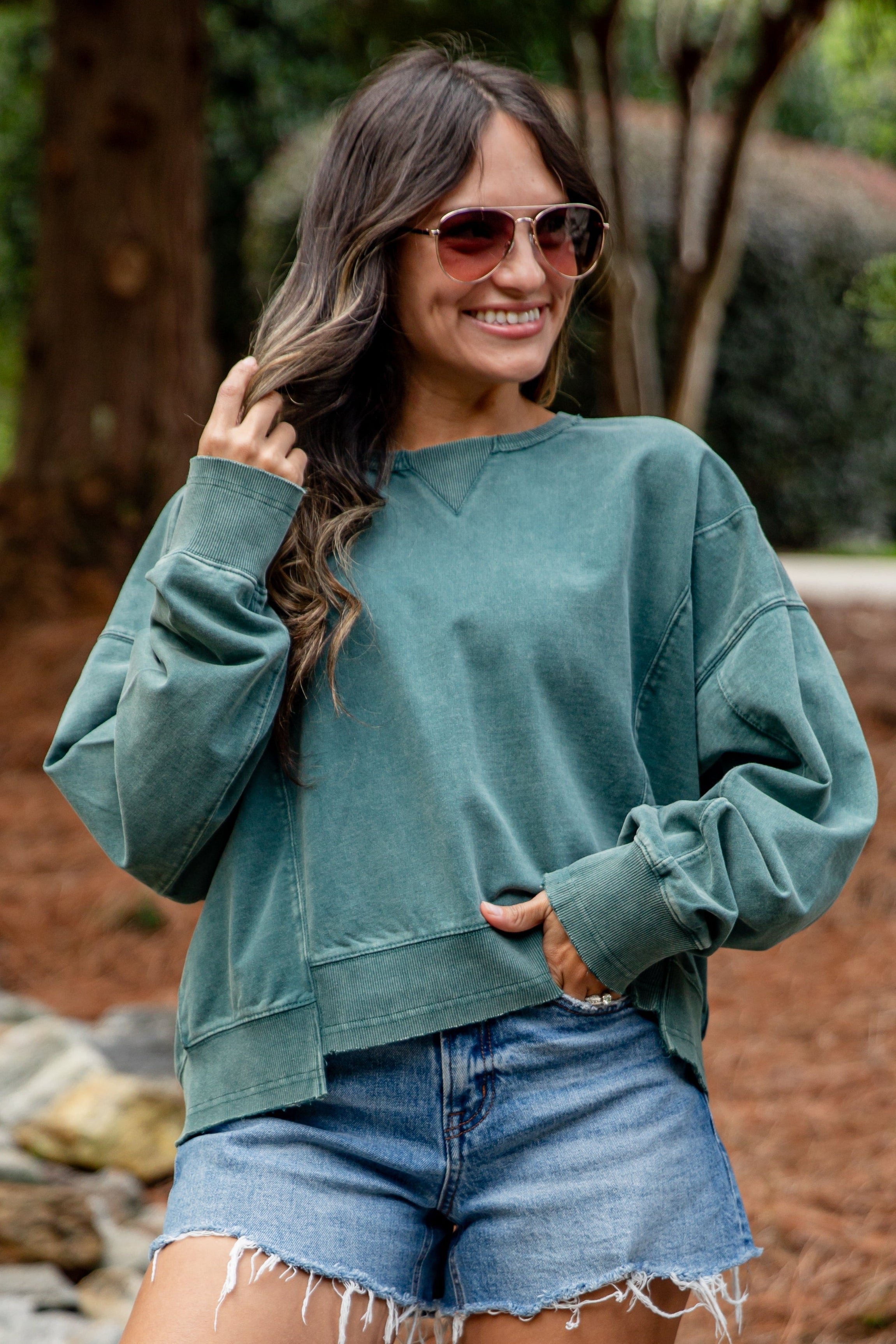 Woman wearing a green sweatshirt and denim shorts standing outdoors with trees in the background