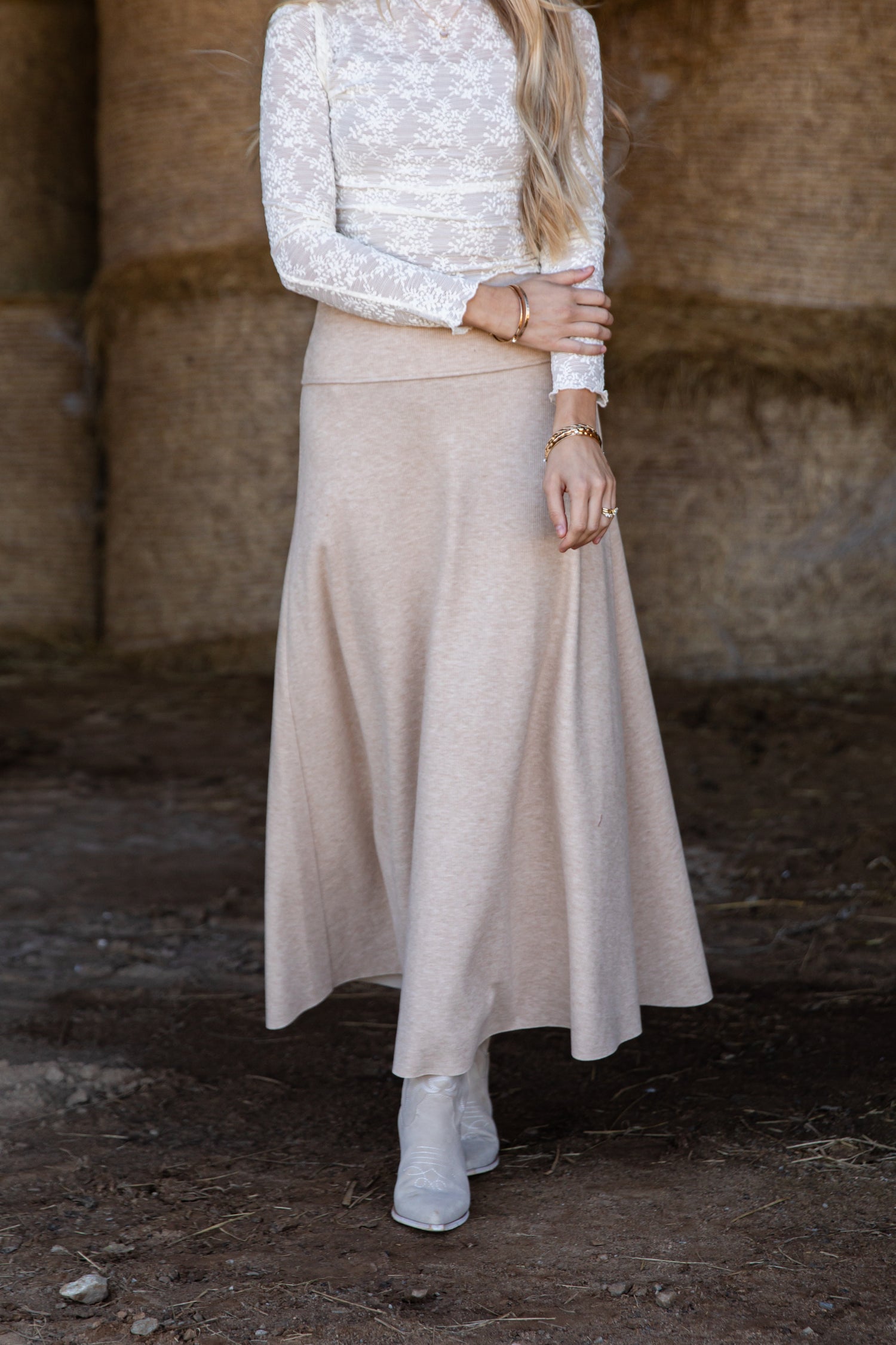 Person wearing a white long-sleeve top and beige skirt standing against a stone wall.