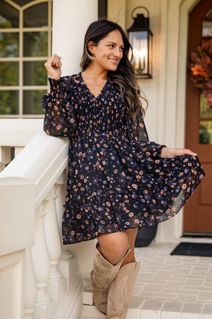 Woman in a floral dress and knee-high boots standing on a porch.