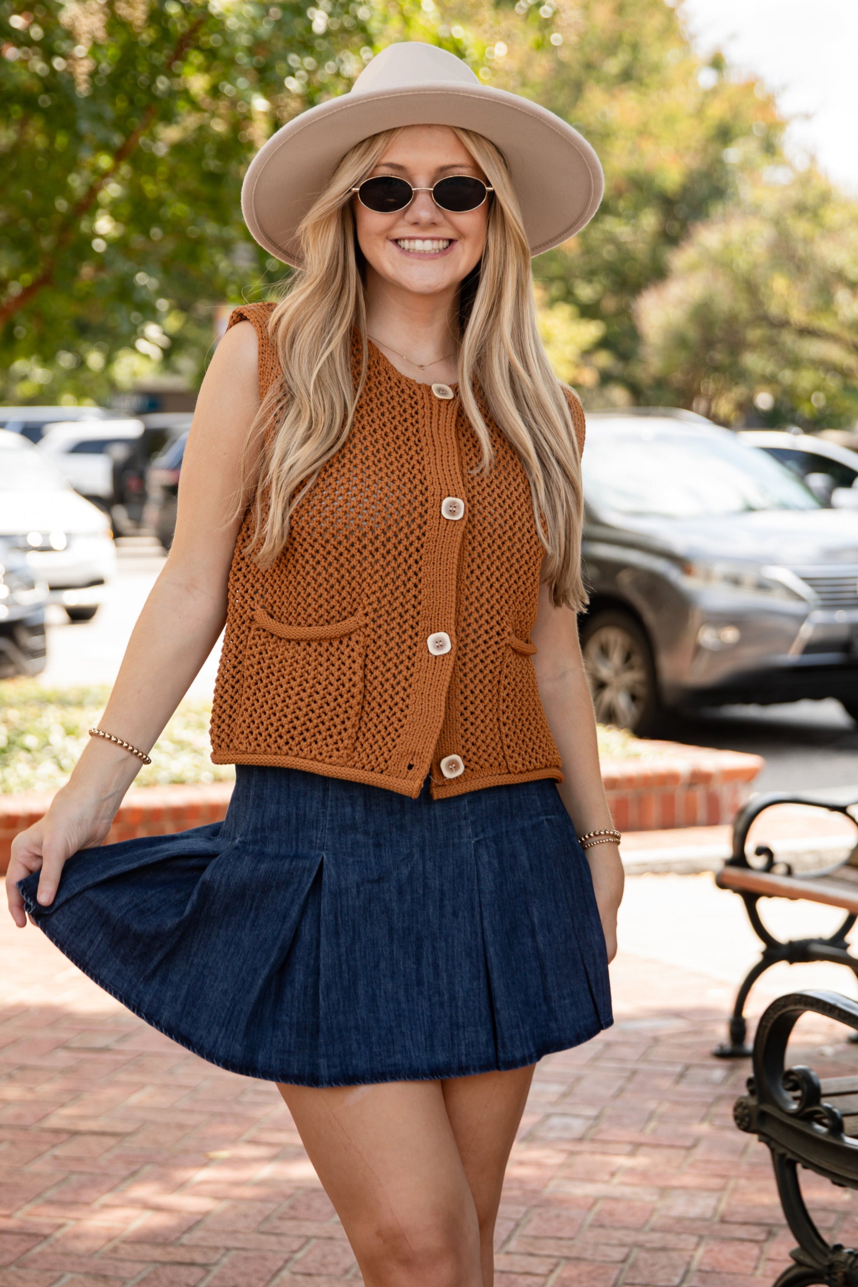 Woman in a brown top and blue skirt standing on a sidewalk with cars and trees in the background.