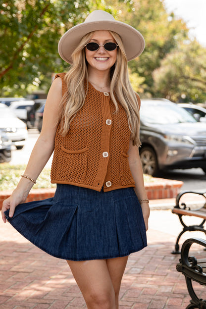 Woman in a brown top and blue skirt standing on a sidewalk with cars and trees in the background.