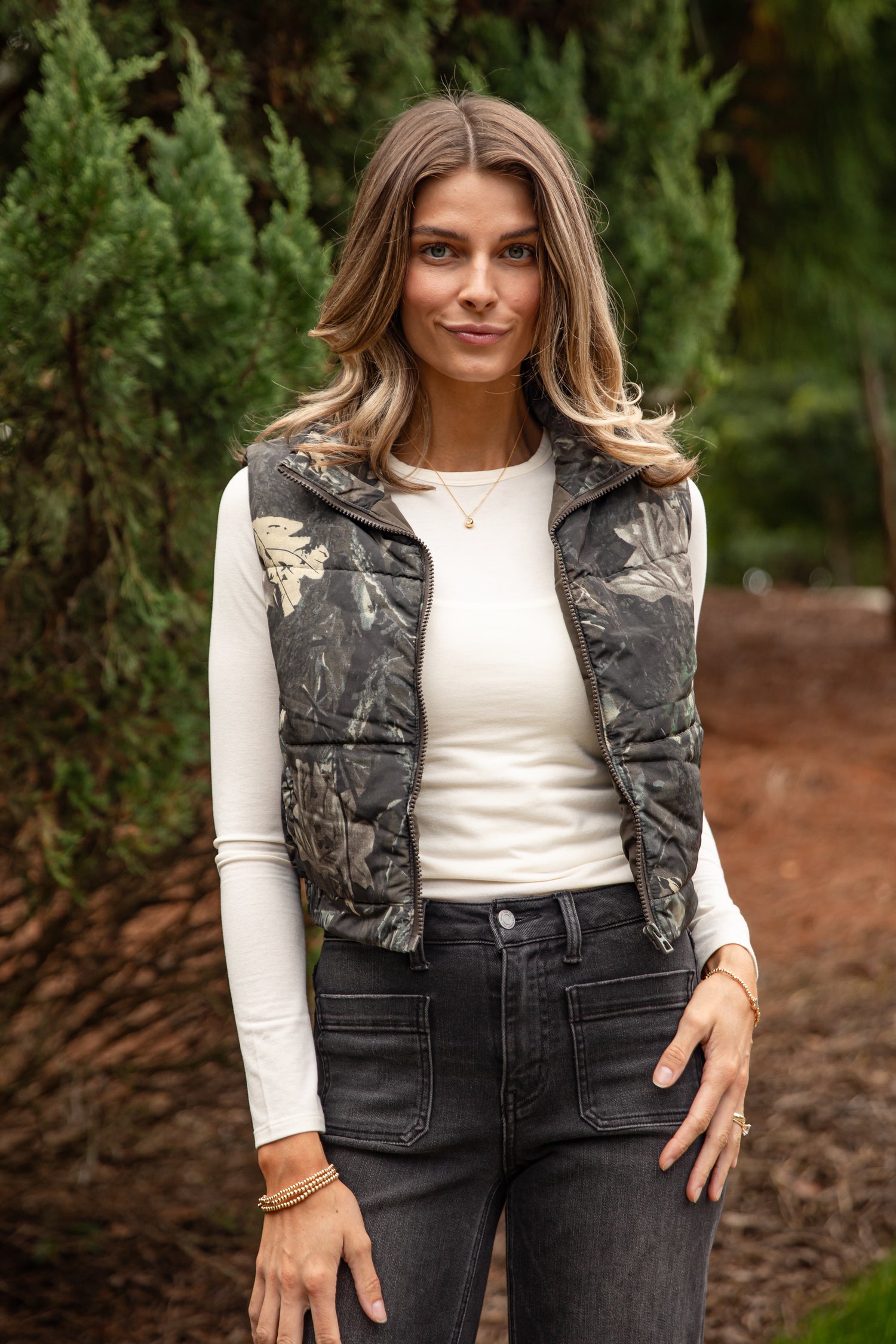 Woman wearing a camouflage vest over a white shirt and dark jeans outdoors.
