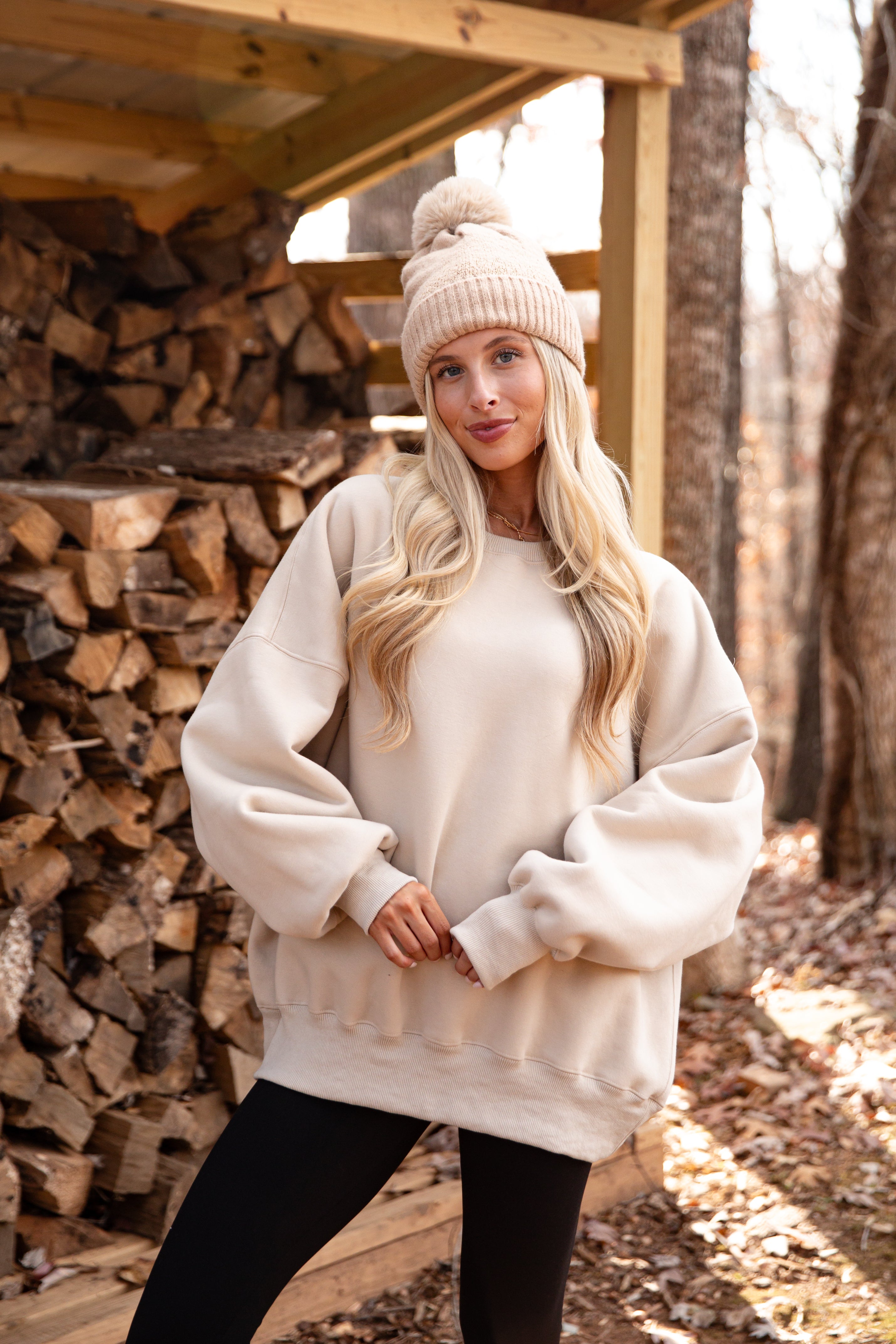 Woman wearing a cream sweater and beige beanie standing in front of stacked firewood.