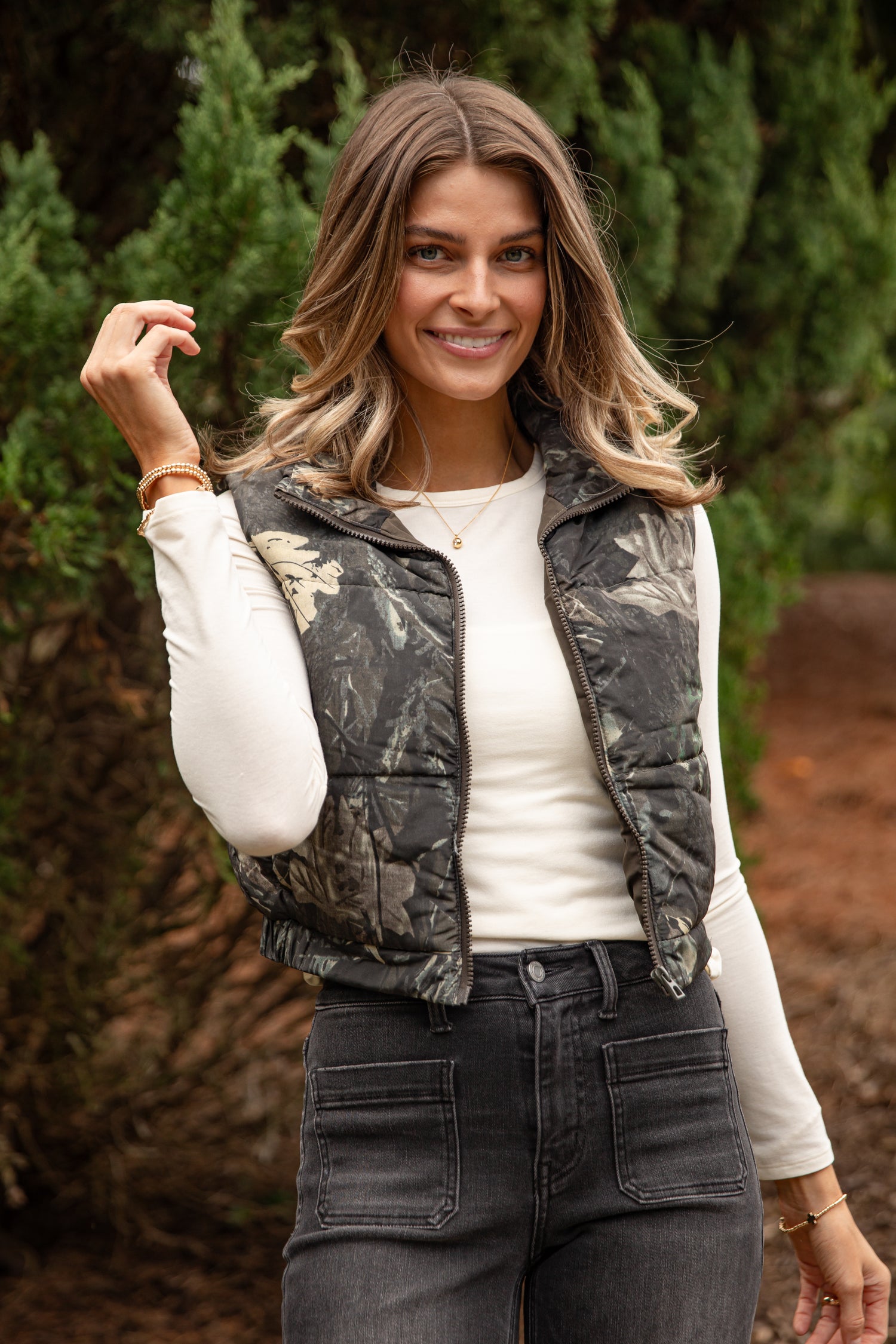 Woman wearing a camouflage vest over a white shirt with trees in the background