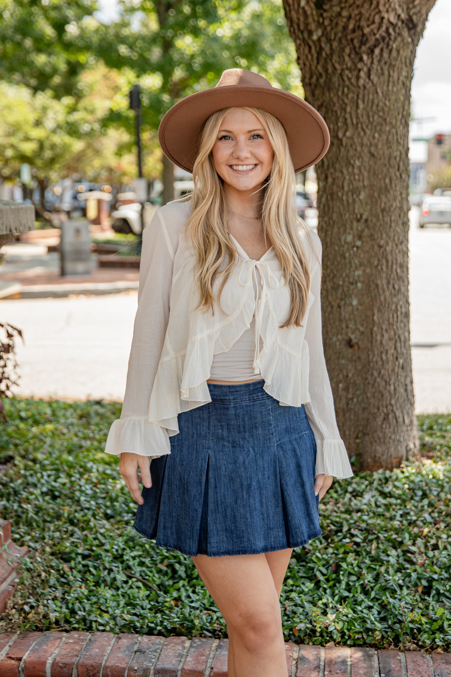 Woman wearing a beige hat, white blouse, and blue skirt standing outdoors.