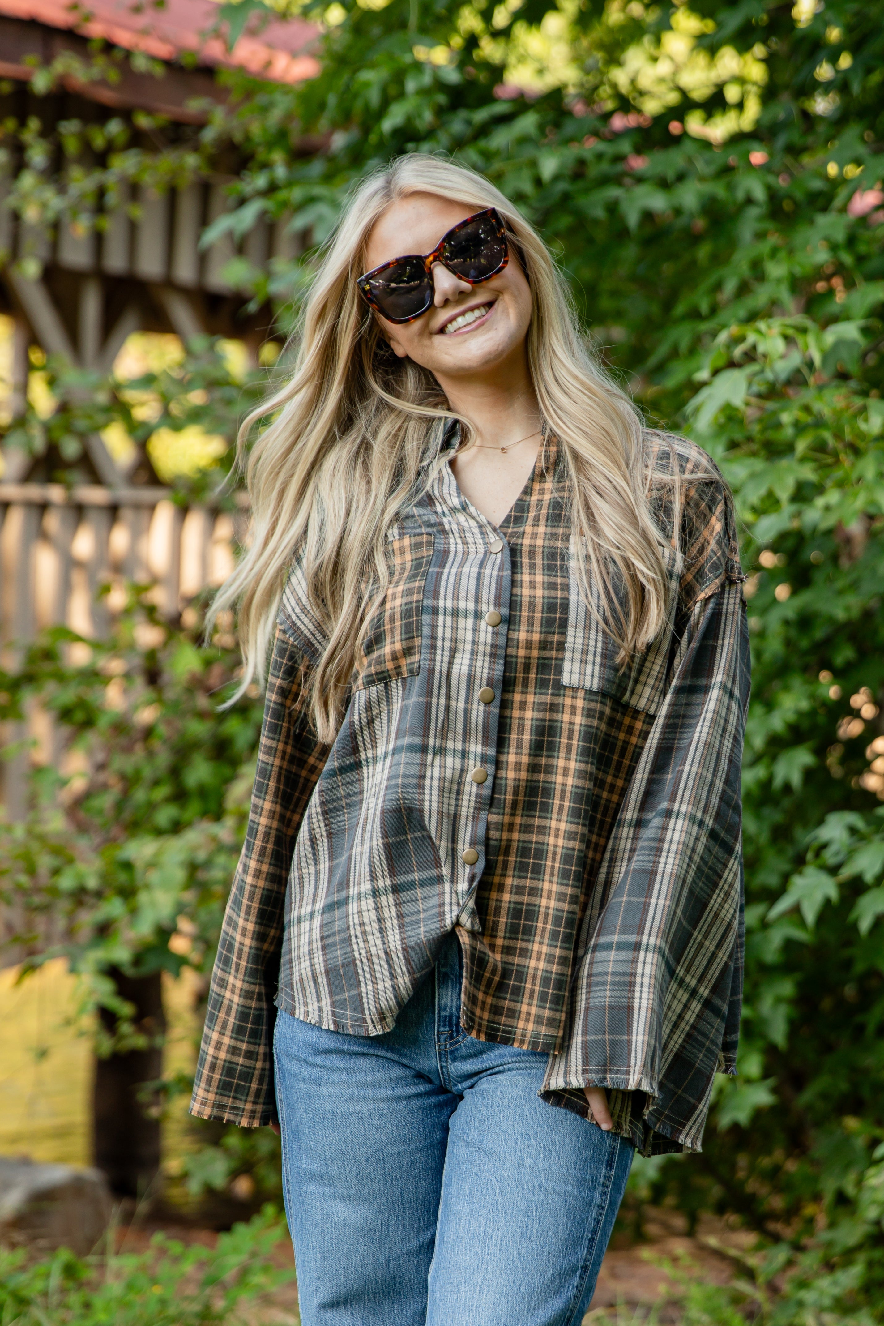 Woman wearing a plaid shirt and sunglasses outdoors with greenery in the background