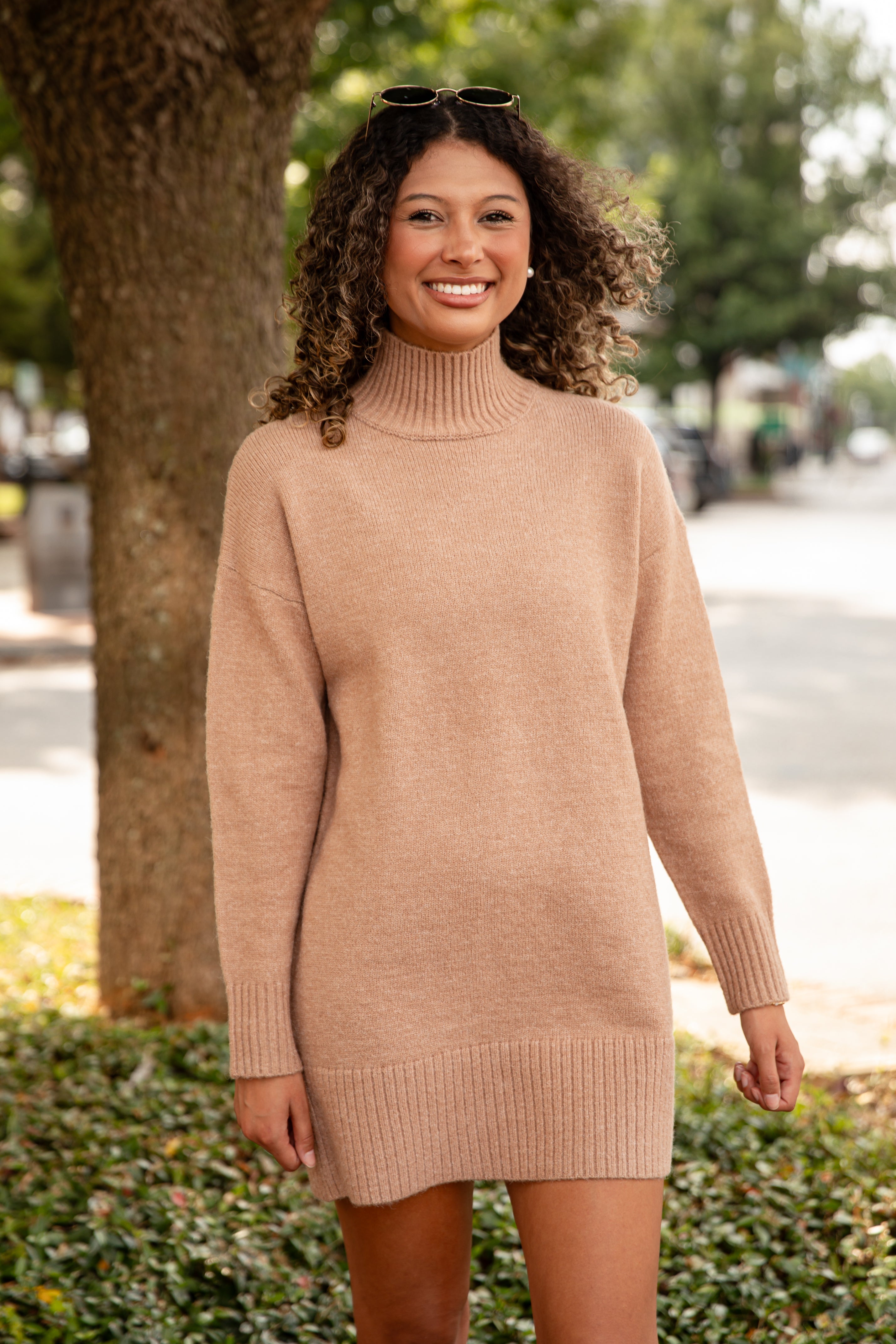 Woman wearing a beige sweater dress standing outdoors with trees and a street in the background.