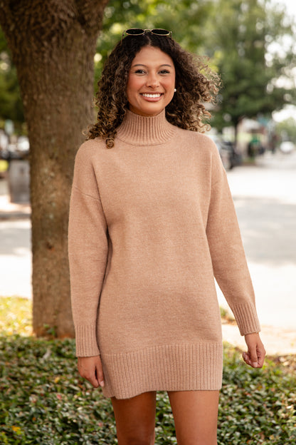 Woman wearing a beige sweater dress standing outdoors with trees and a street in the background.