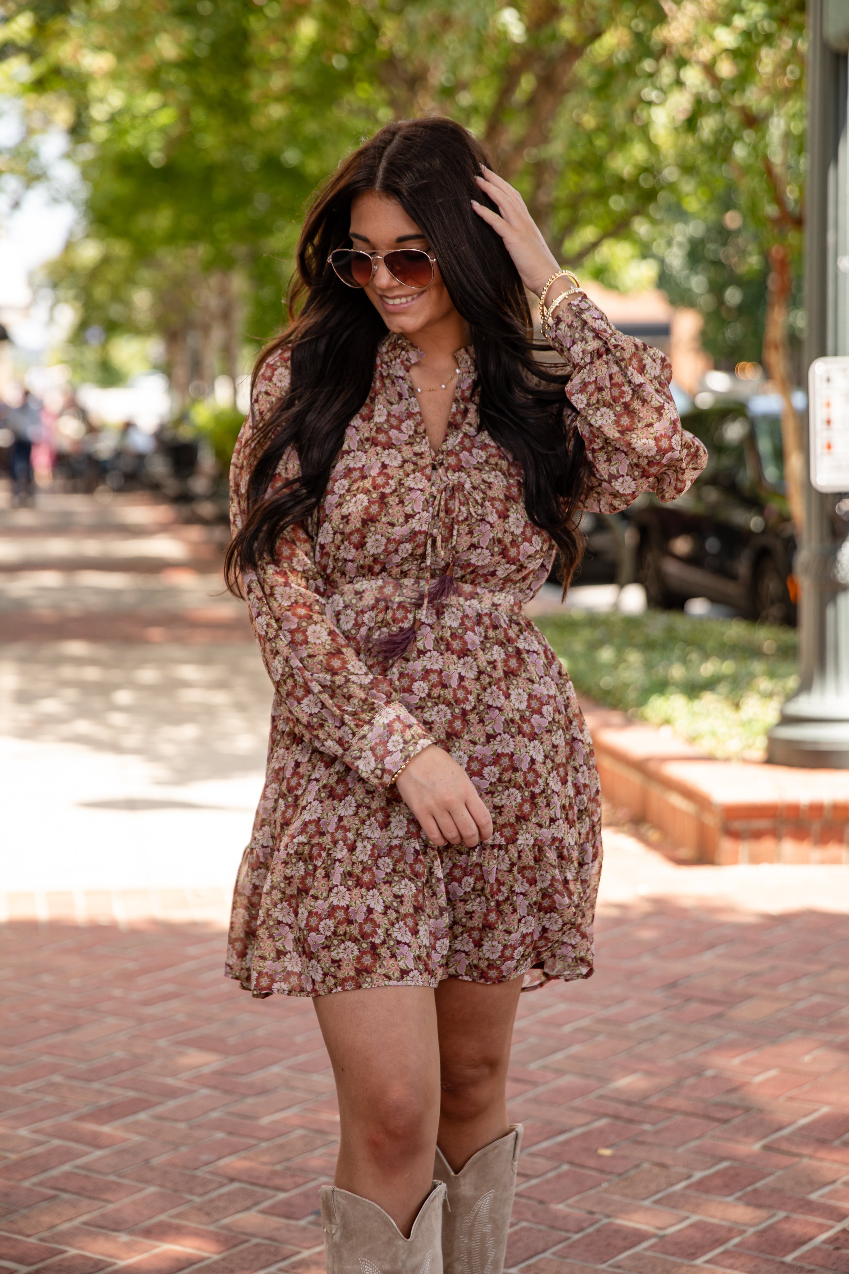 Woman in a floral dress and boots standing on a sidewalk with trees in the background