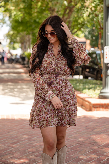 Woman in a floral dress and boots standing on a sidewalk with trees in the background