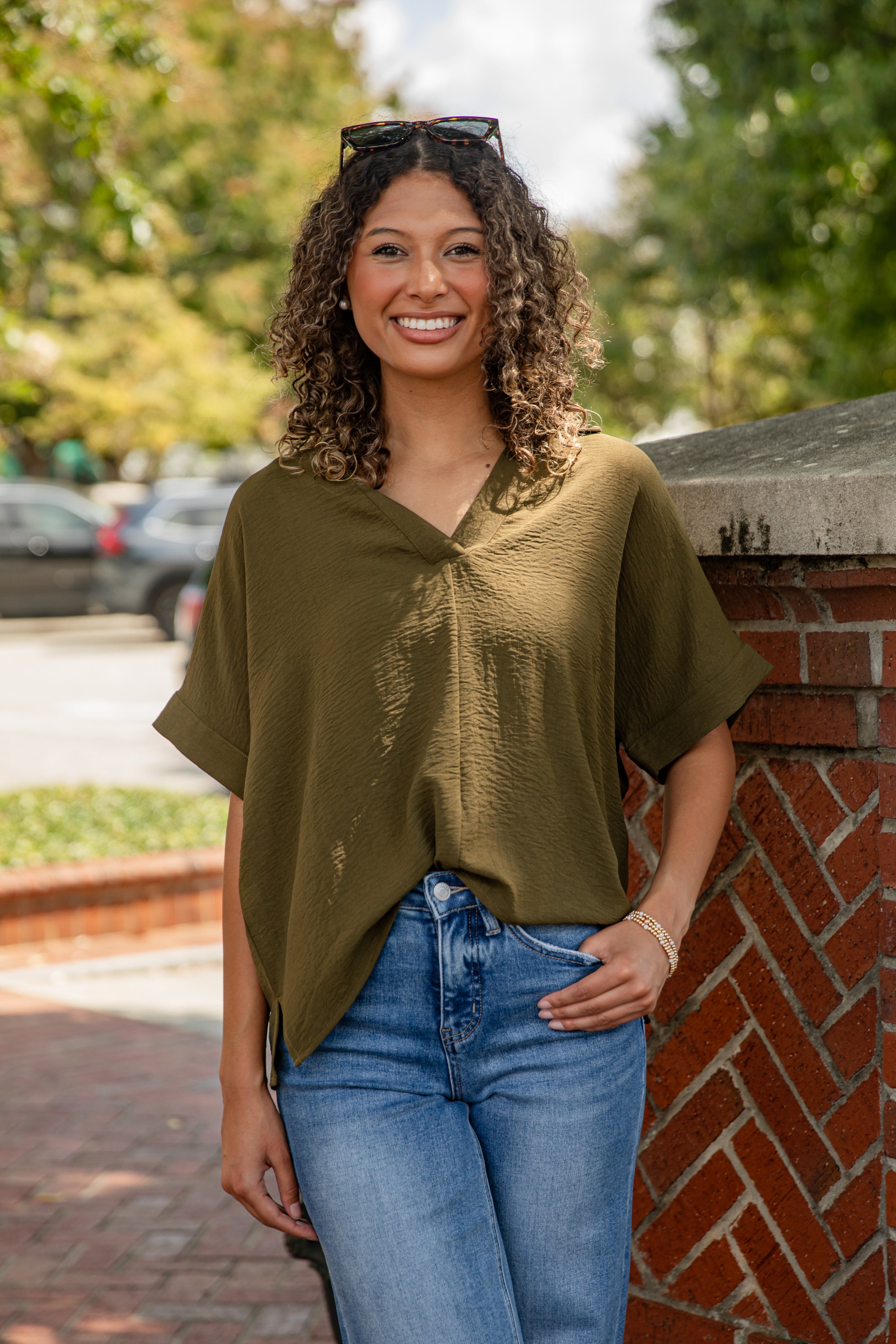Woman wearing an olive green top and blue jeans standing against a brick wall with trees in the background.