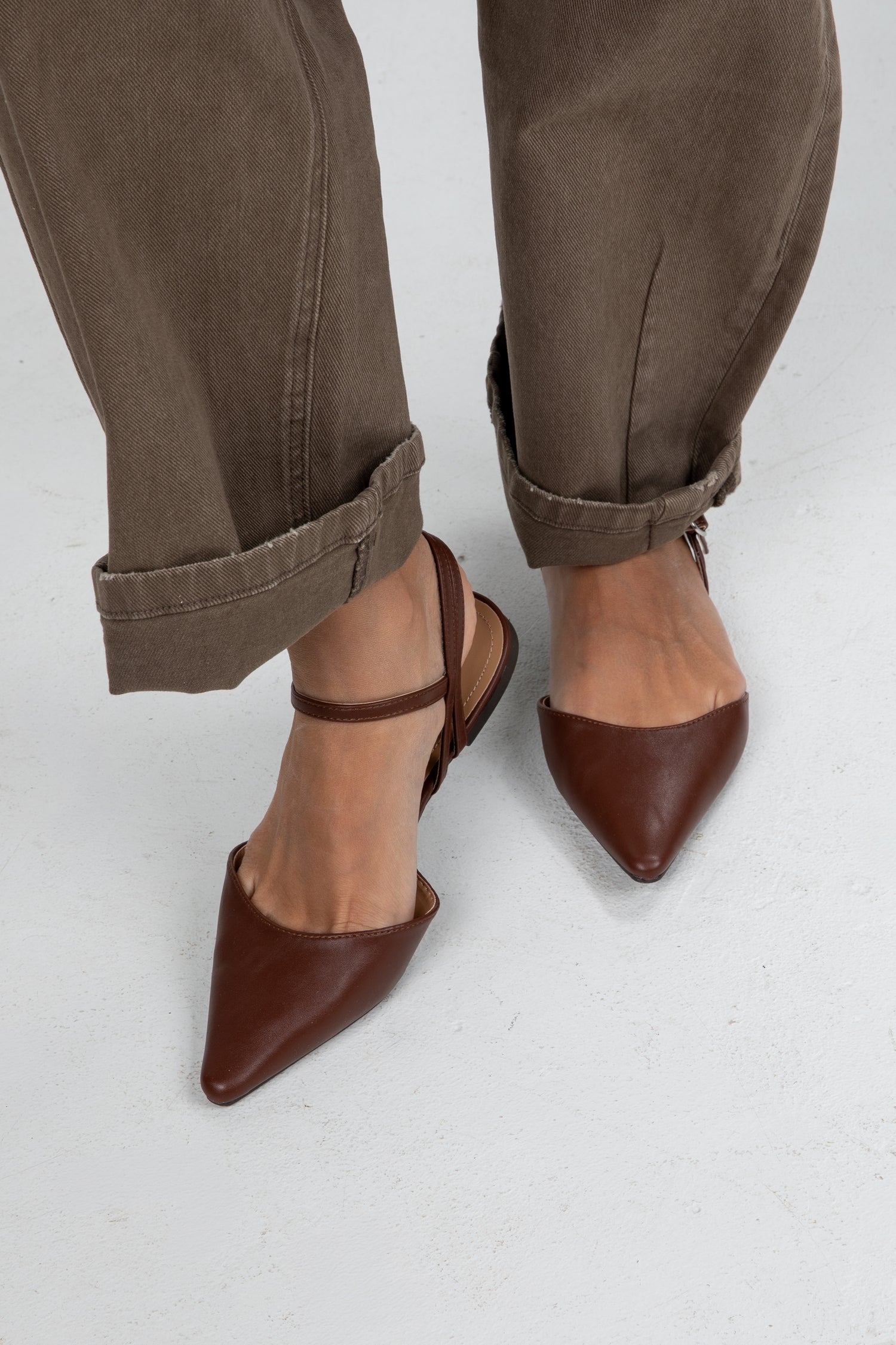 Brown pointed-toe shoes worn with rolled-up brown pants on a light gray background