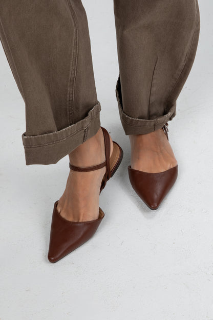Brown pointed-toe shoes worn with rolled-up brown pants on a light gray background