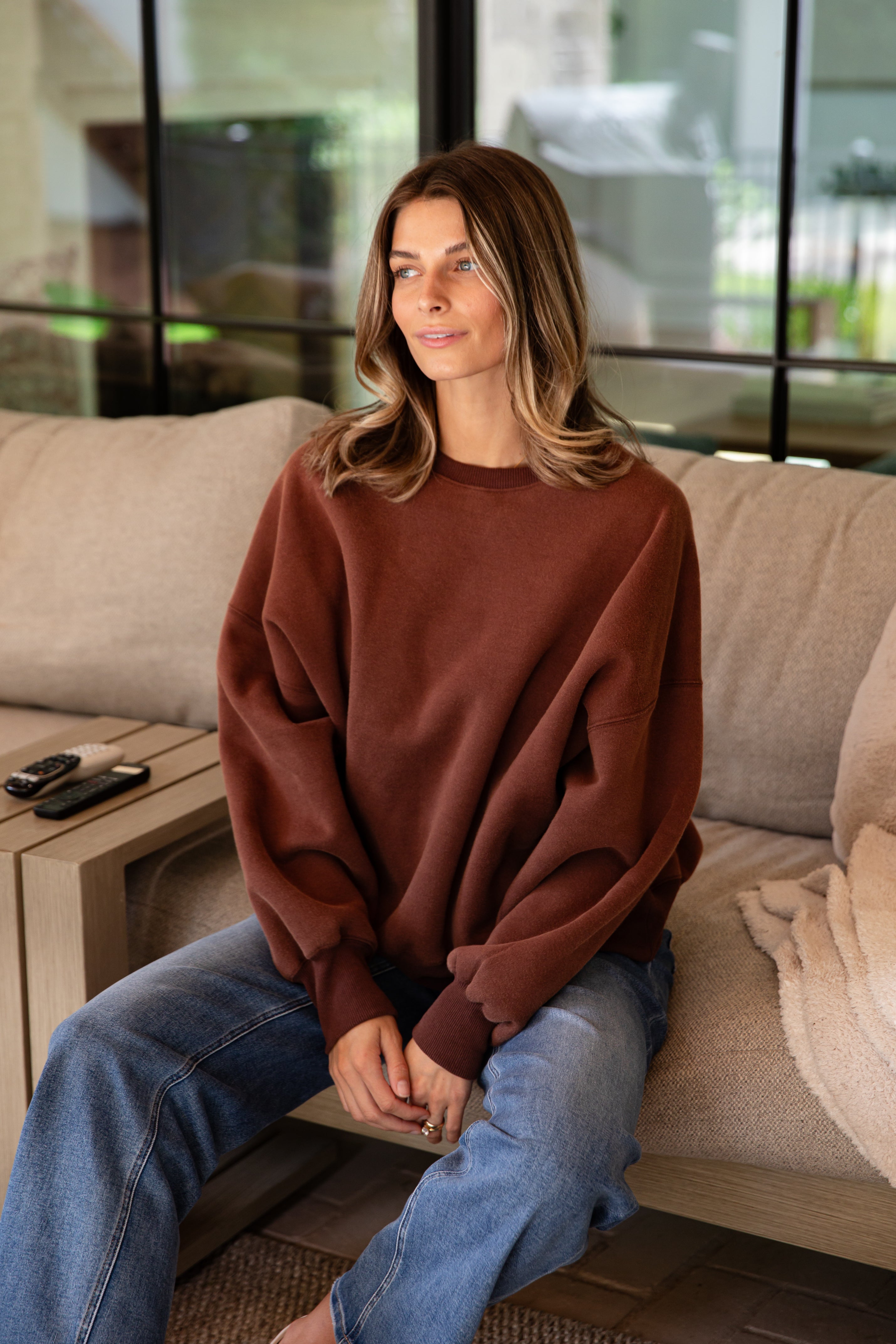 Woman sitting on a couch wearing a brown sweater and blue jeans in a modern living room.