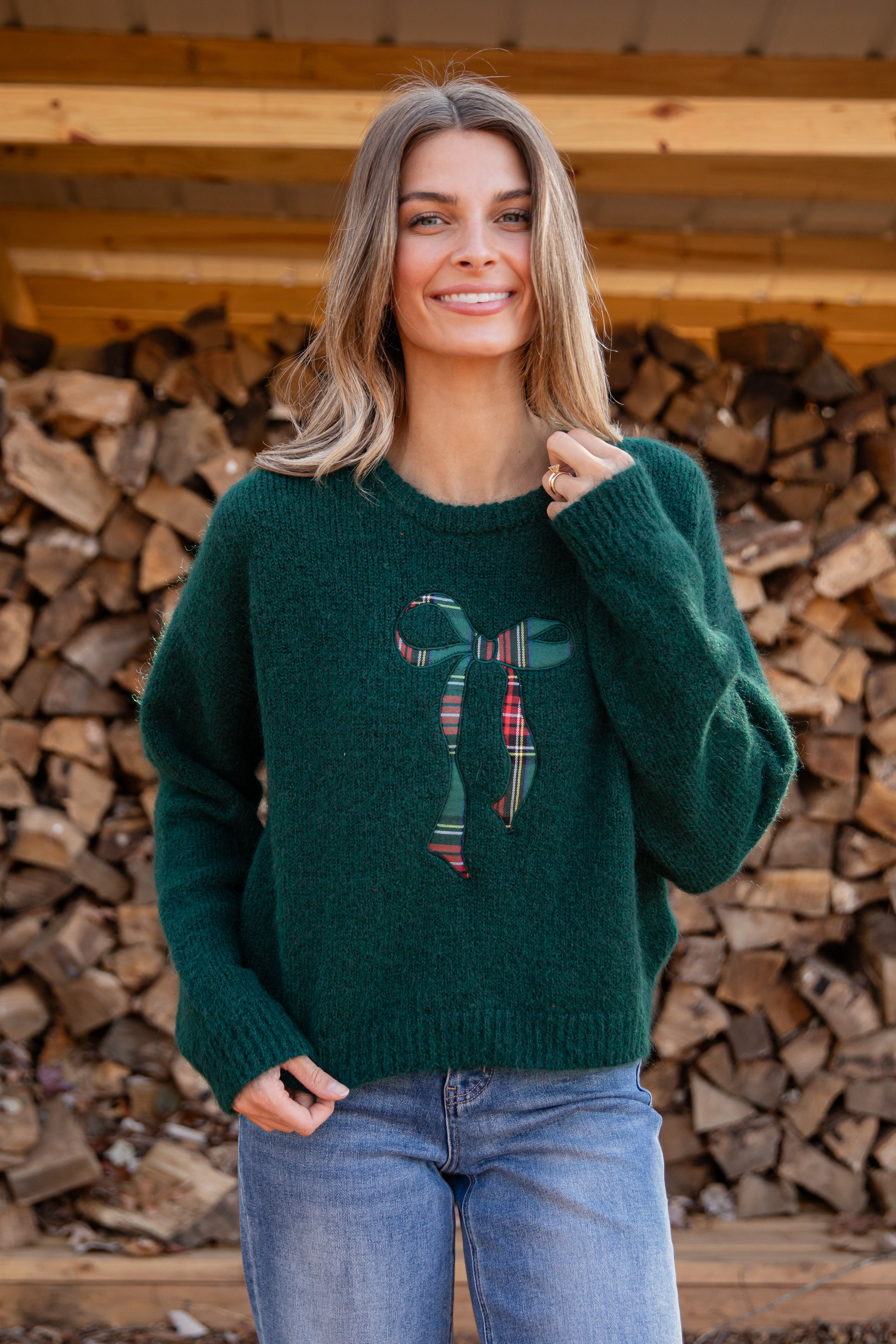 Woman wearing a green sweater with a bow design in front of stacked firewood.