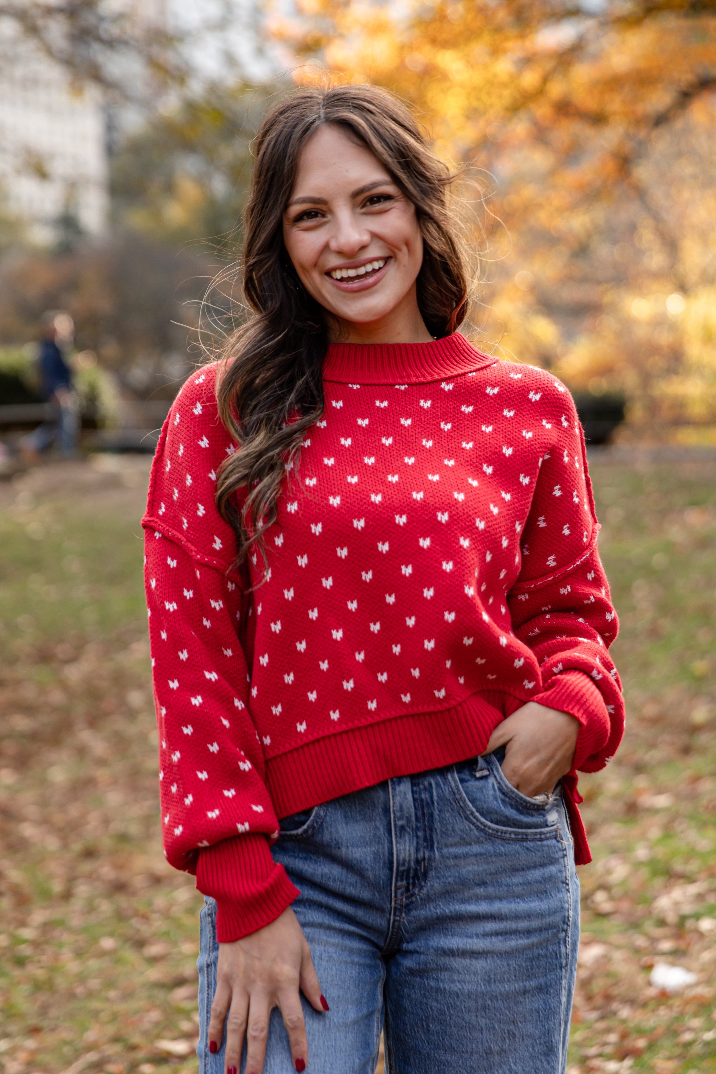 Woman wearing a red sweater with white patterns and blue jeans standing in an outdoor setting with trees and people in the background.