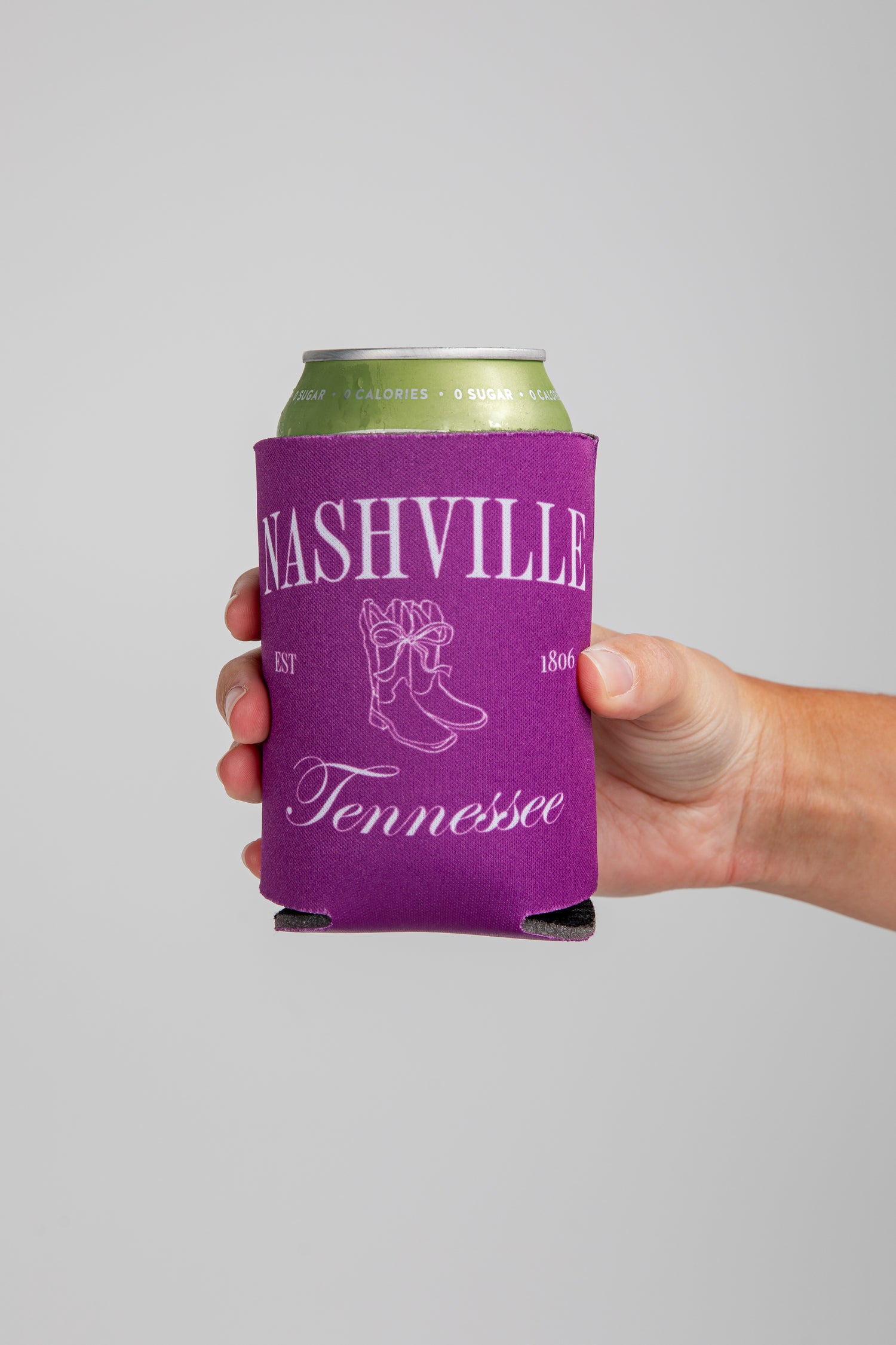 Hand holding a can with a purple Nashville, Tennessee koozie against a gray background