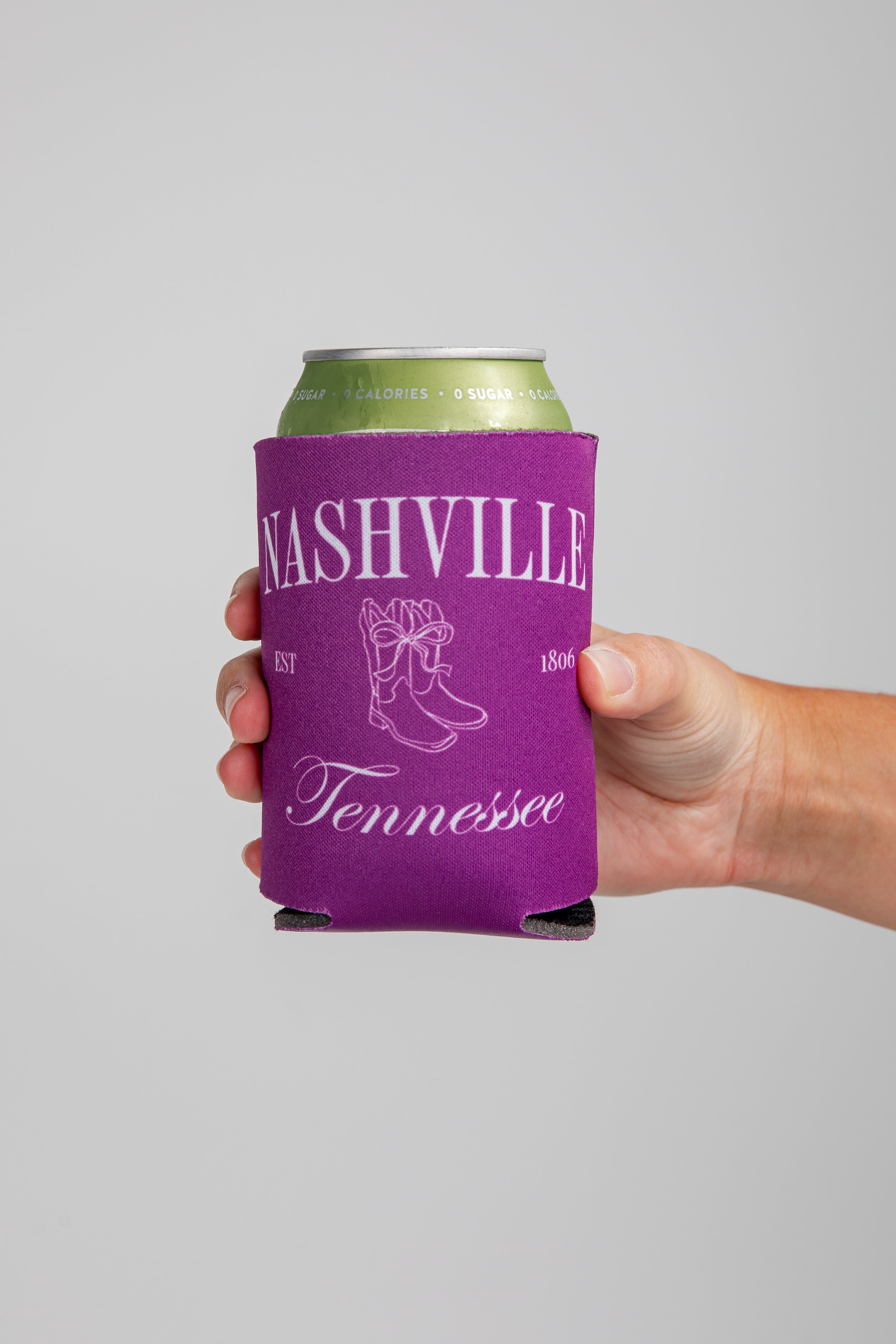 Hand holding a can with a purple Nashville, Tennessee koozie against a gray background
