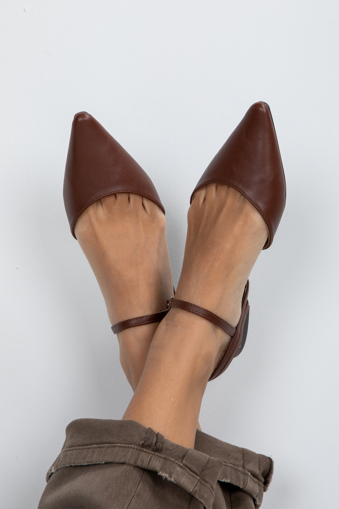 Brown pointed-toe shoes with ankle straps worn by a person on a white background