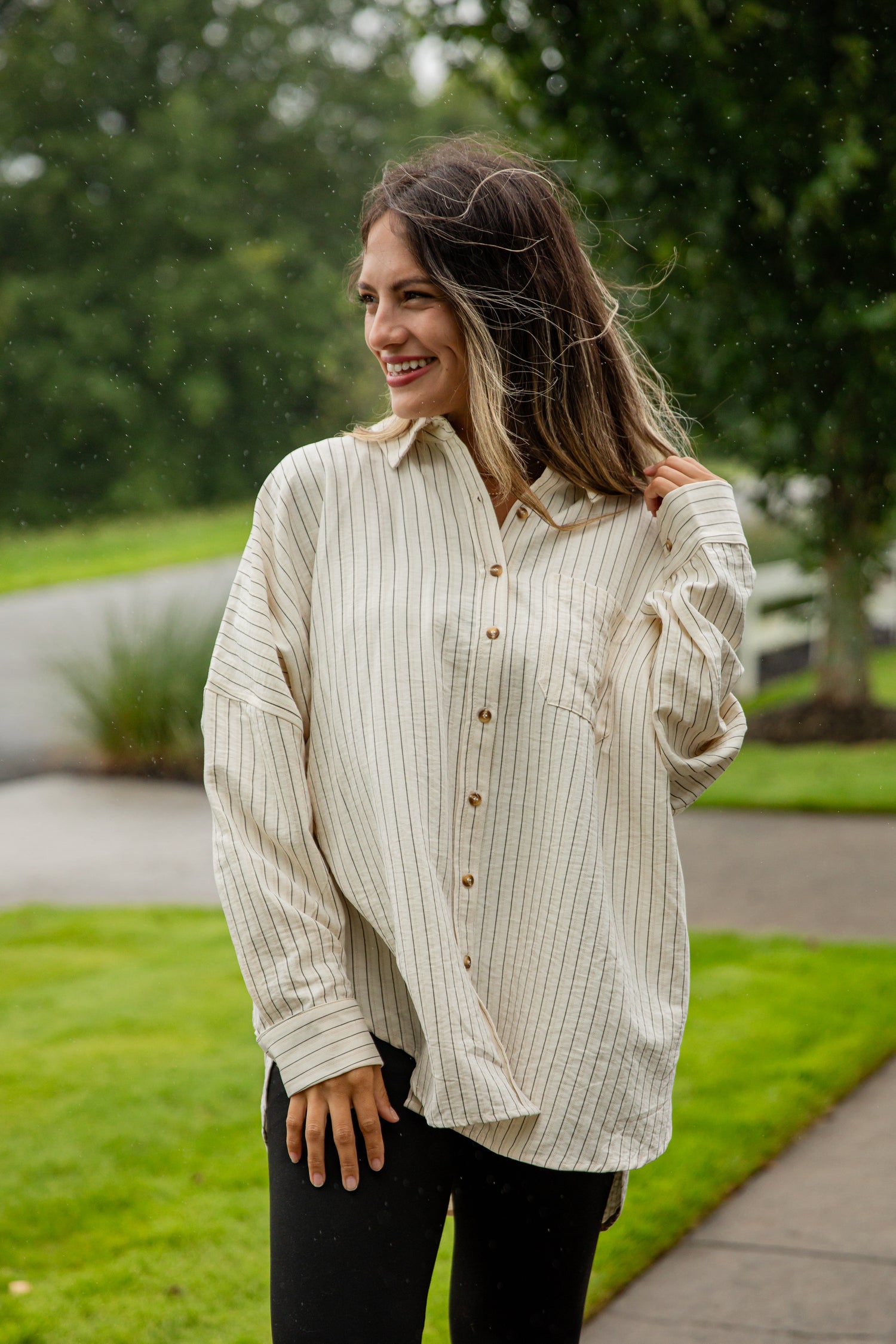 Woman wearing a light-colored button-up shirt outdoors with greenery in the background