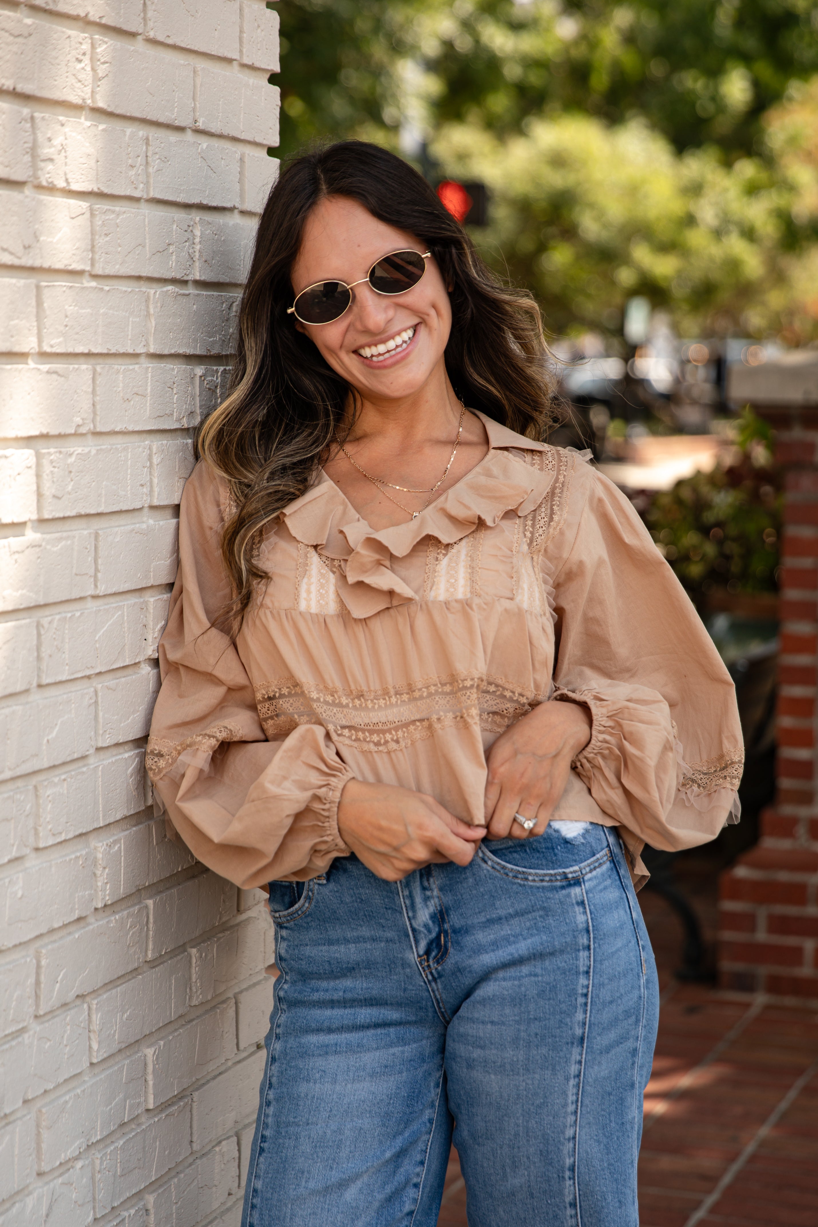 Woman wearing a beige blouse and blue jeans leaning against a brick wall.