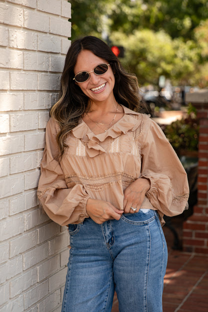 Woman wearing a beige blouse and blue jeans leaning against a brick wall.