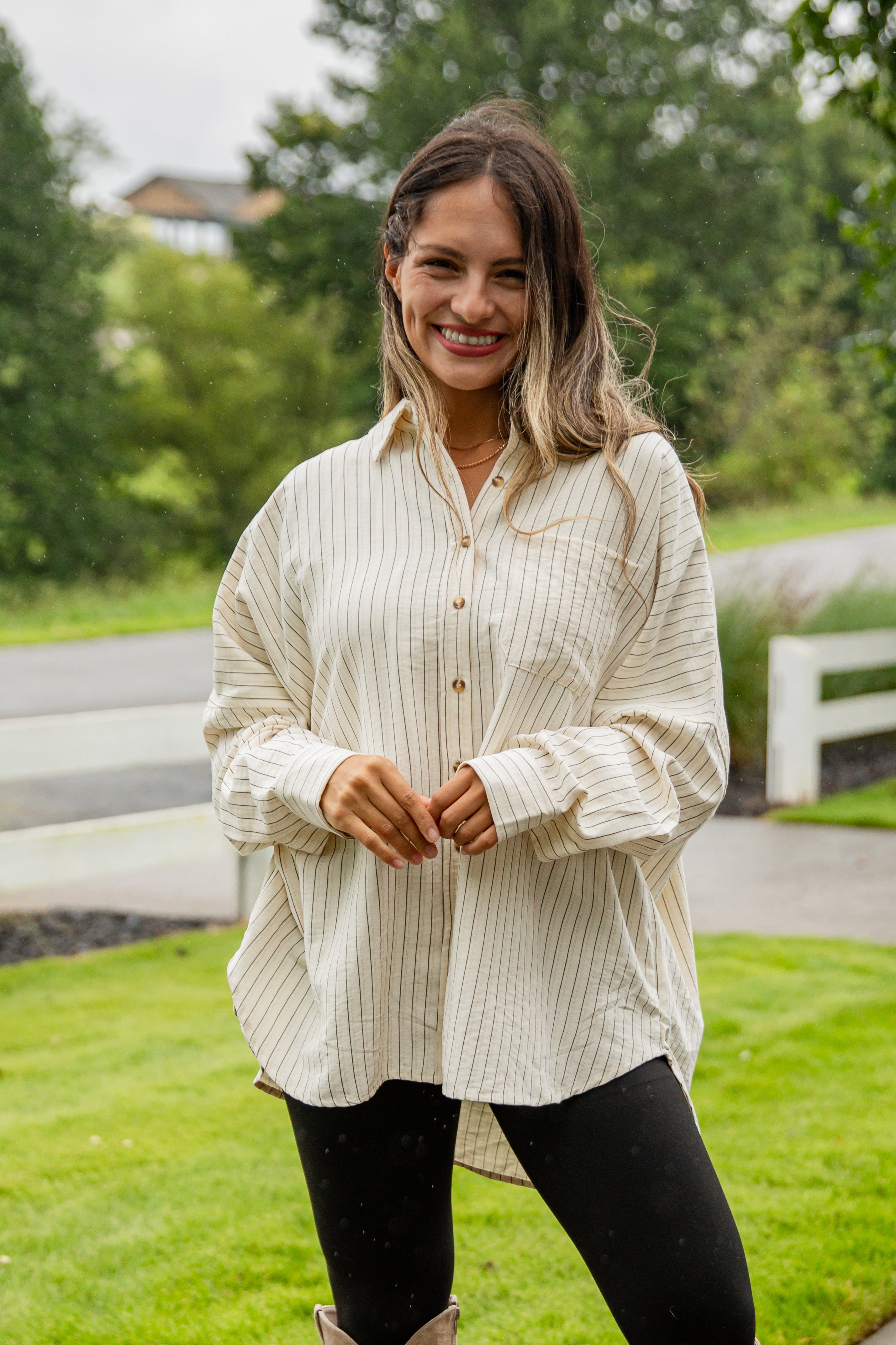 Woman wearing a light-colored button-up shirt and dark pants standing outdoors with greenery in the background