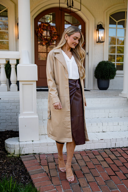 Woman in a beige trench coat and brown leather skirt standing on a porch.
