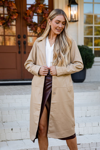 Woman wearing a beige trench coat standing outside a house.
