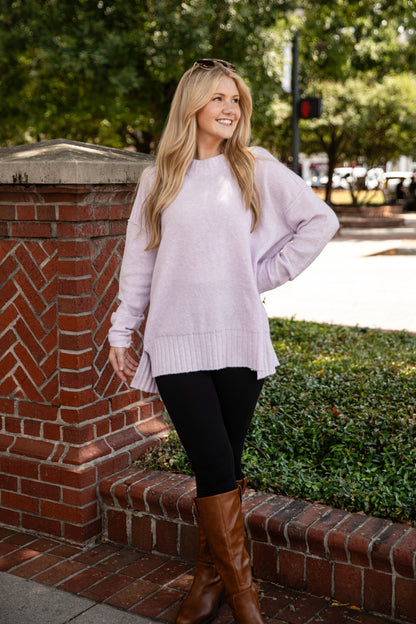 Woman wearing a light purple sweater and brown boots standing on a sidewalk with greenery and a brick wall in the background.